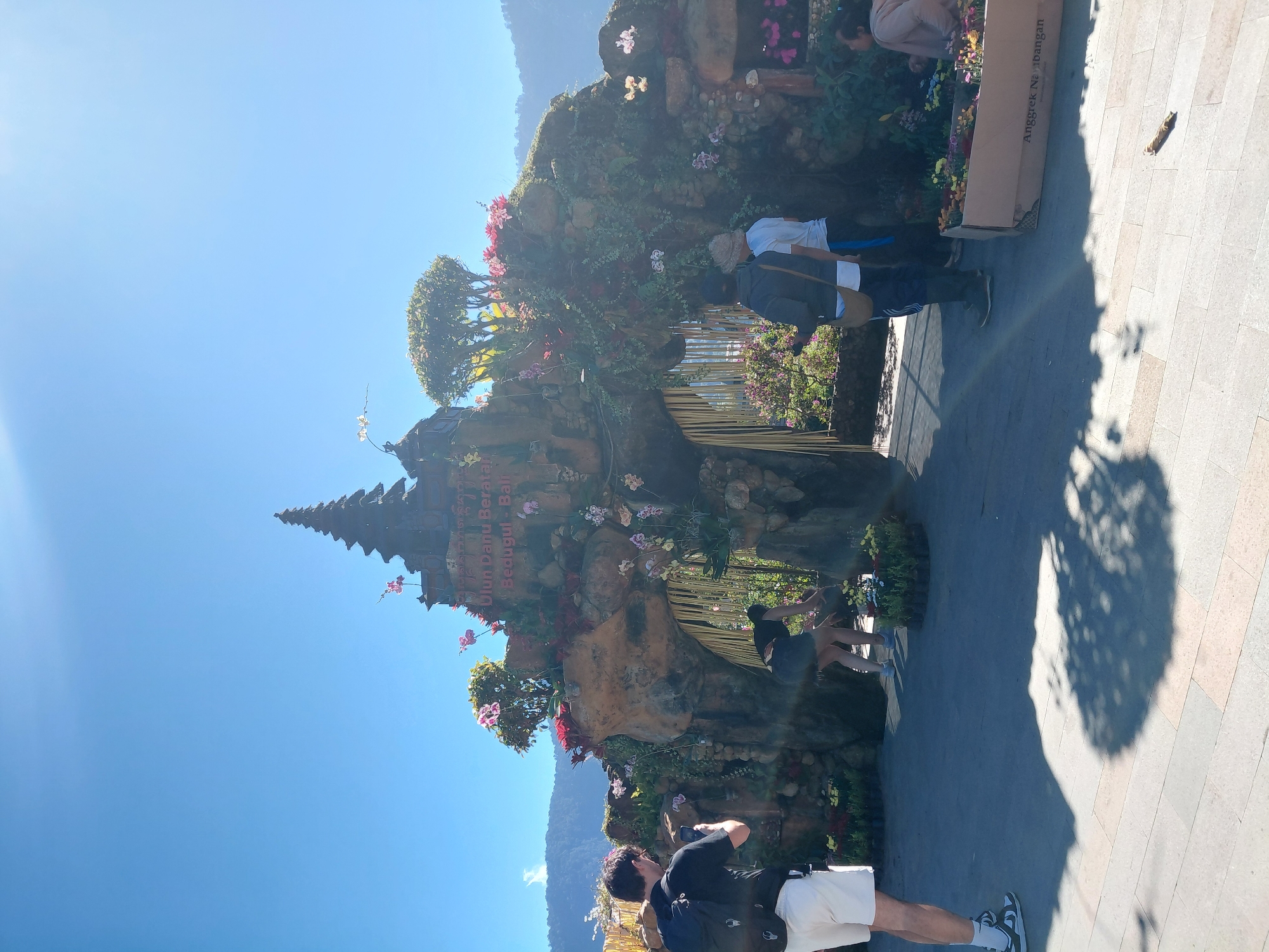 The image shows the entrance to the Ulun Danu Beratan Temple in Bedugul, Bali, Indonesia. The entrance is a large, ornate structure decorated with flowers and greenery, designed to resemble a stone archway. The temple's name is visible in text on the structure. Several people are present, with some posing for photos and others engaged in activities. One person, wearing a black t-shirt and white shorts, is taking a photo with their phone. Another person in a black top and skirt is seen bending down, possibly tending to plants. Two people in the background are facing the entrance, and another person is seated on the right side of the frame. The environment is outdoors, with a clear blue sky and mountains visible in the distance. The ground is paved with light-colored stone tiles, and shadows from trees are cast on the ground. There are also planters filled with flowers, and a cardboard box with text on it.