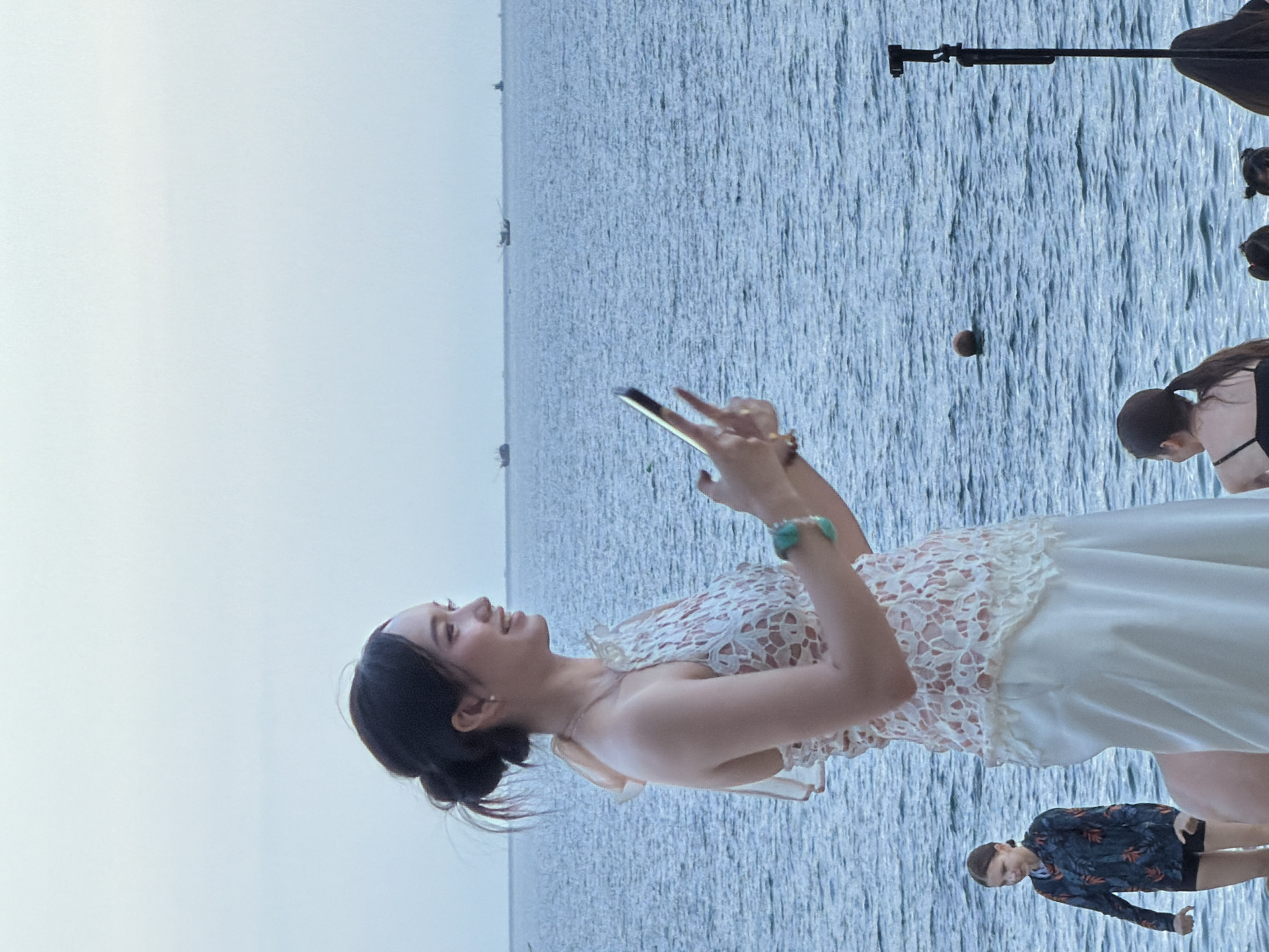 A woman is holding a smartphone and taking a picture with a beach and the ocean in the background. She is wearing a white dress with lace detail. There are several boats in the distance on the ocean. A young girl in a patterned rash guard and shorts is standing to the left of the woman. The scene appears to be on a beach in Nha Trang, Vietnam.