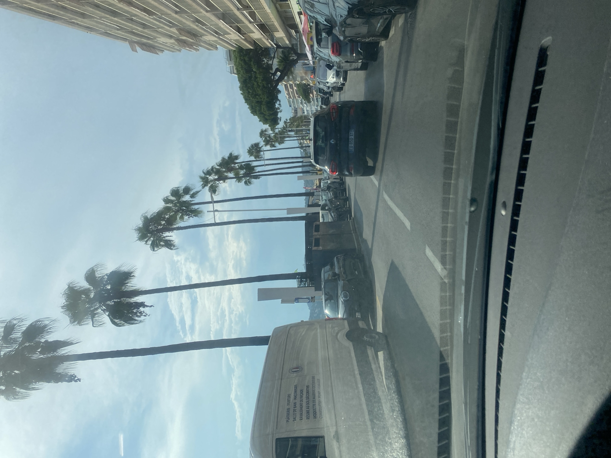 The image depicts a street scene in Antibes, France, likely along the coast. A line of tall palm trees lines the left side of the road, with a body of water visible beyond them. On the right side of the road, a multi-story building with balconies is prominent. Several cars are visible, driving in traffic. In the foreground, a white van with writing on its side, possibly a service vehicle, is parked. Next to it, a black Mini Cooper is also parked. Further down the road, more vehicles are seen, including a black SUV with a license plate that reads "EG 650 XY". The sky is partly cloudy.