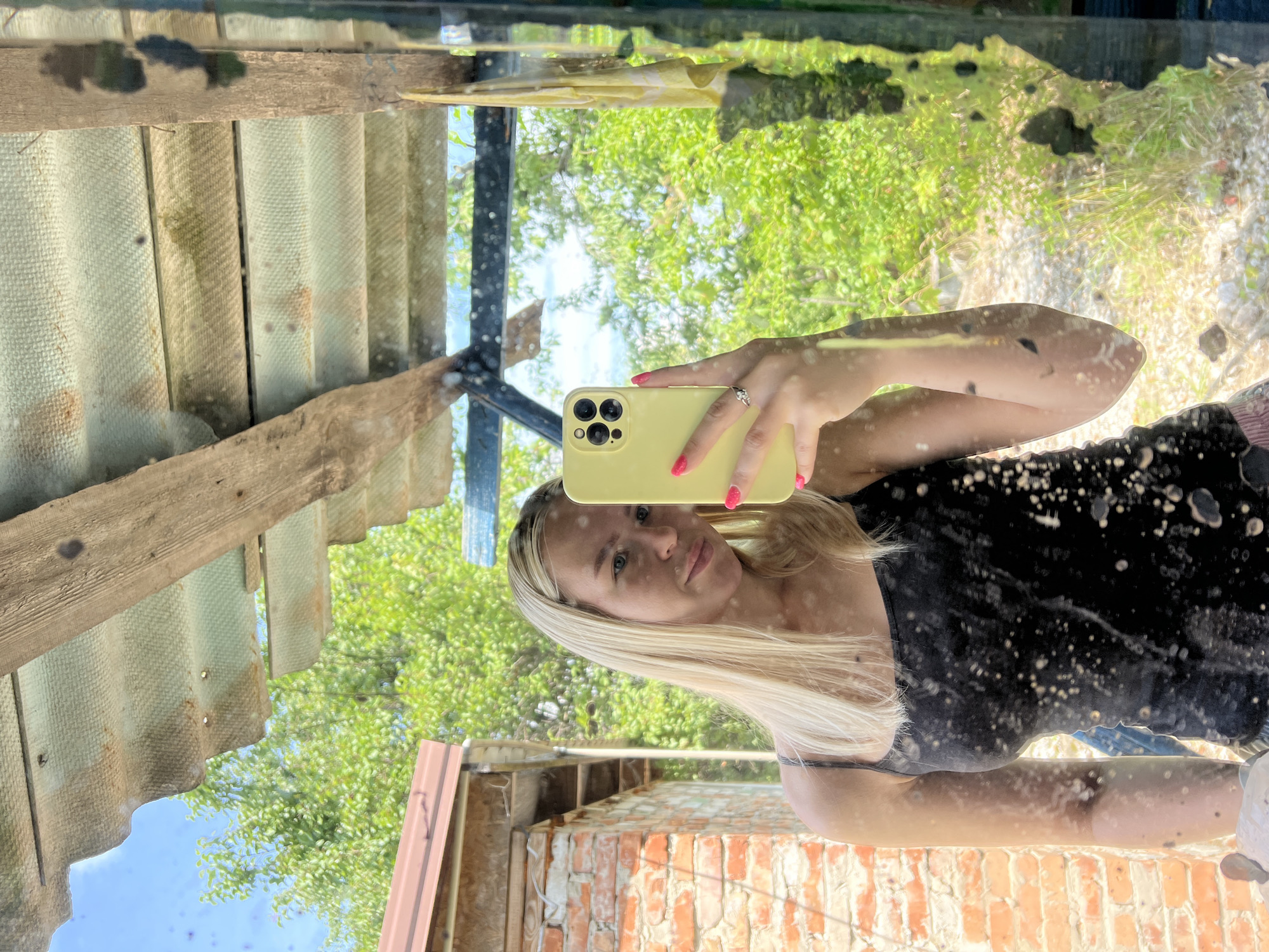 A woman with blonde hair is taking a selfie in a mirror. She is wearing a black tank top and a ring on her left hand. She is holding a yellow smartphone in her right hand. The background appears to be outdoors, with trees and a brick building visible. The mirror is dirty and smudged, with many spots on its surface. Above the woman, there is a rustic roof structure made of wood and corrugated metal sheets. The scene is bathed in natural light, suggesting it is daytime.