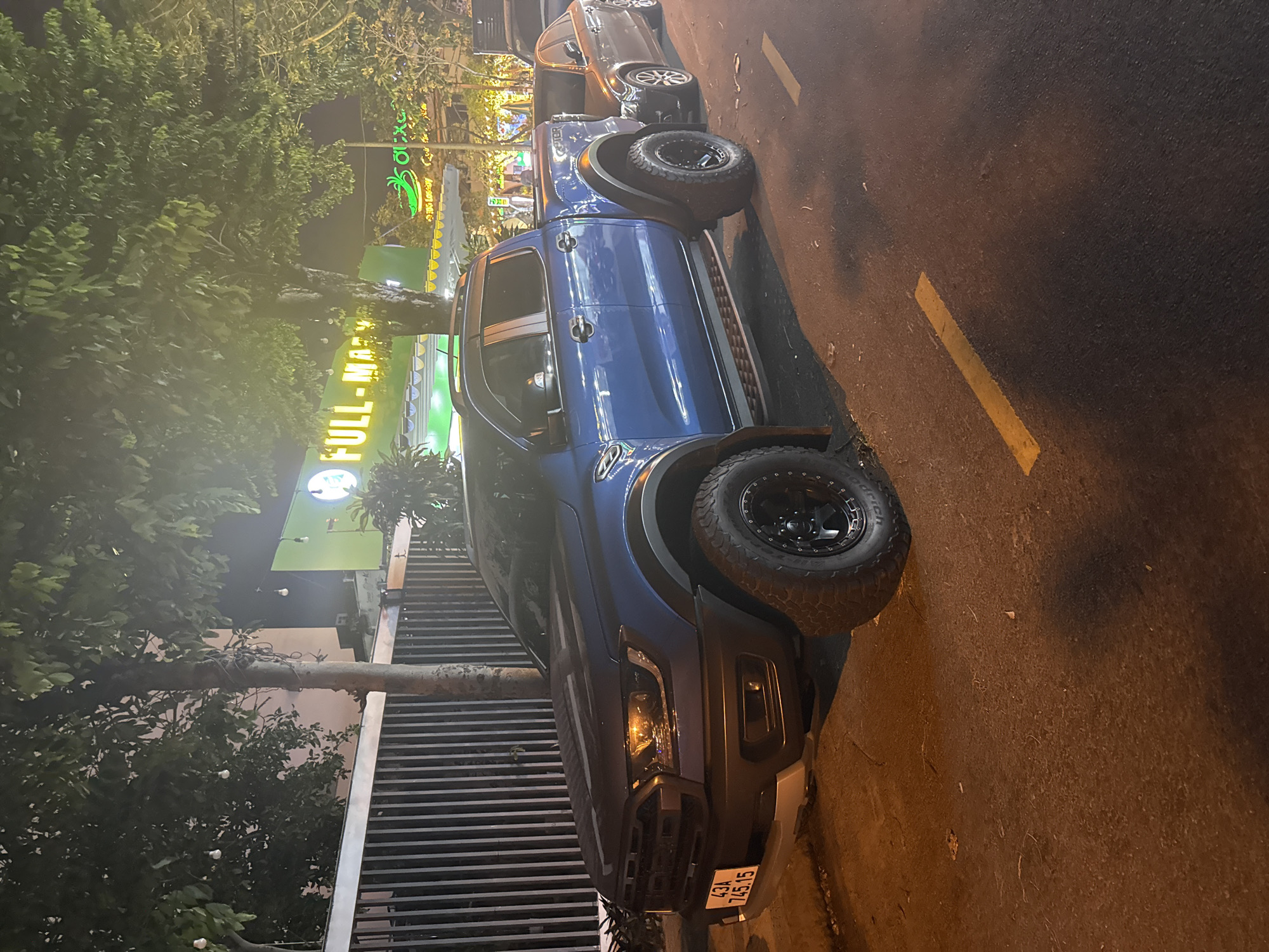 This night-time image, taken in Đà Nẵng, Vietnam, features a blue Ford Ranger pickup truck prominently parked on the side of an urban street. The truck is equipped with rugged, knobby off-road tires on black rims, and its front license plate, displaying "43A 745.15," is clearly visible. Behind the blue truck, another dark-colored sedan or SUV is partially visible.
The setting is an urban environment illuminated by streetlights, casting warm, yellowish light on the dark asphalt road, which has faint yellow markings. Adjacent to the parked vehicles is a building with bright, glowing signage. One sign, in yellow-green, partially reads "FULL-MA," indicative of a commercial establishment. Above it, another green sign displays an icon resembling a banana or plantain, along with some Vietnamese characters. Lush green trees and foliage are abundant, particularly on the left, partially obscuring the buildings and adding to the street's character. A metal fence or railing runs alongside the building. There are no people visible in the image. The scene suggests a quiet moment on a commercial street after dark.