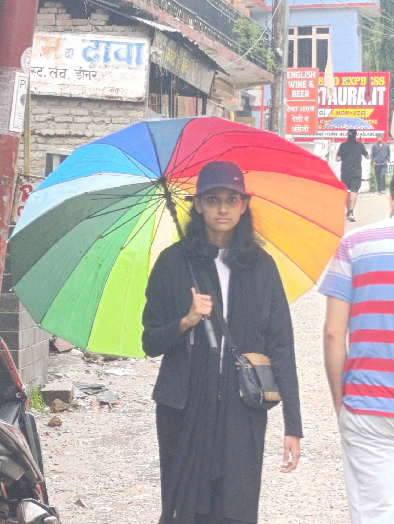 The image shows a street scene in Aleo, India. A young person wearing a black jacket and a baseball cap is holding a large, colorful umbrella, obscuring much of their body. The umbrella has segments of red, orange, yellow, green, and blue. To the right of this person, another individual wearing a striped red, white, and blue shirt and light-colored pants is visible from the chest down. In the background, a building with signage in Hindi and English can be seen, including "ENGLISH WINE & BEER" and "FOOD EXPRESS RESTAURANT." The street is paved with gravel and debris. A motorcycle is partially visible on the left side of the frame.