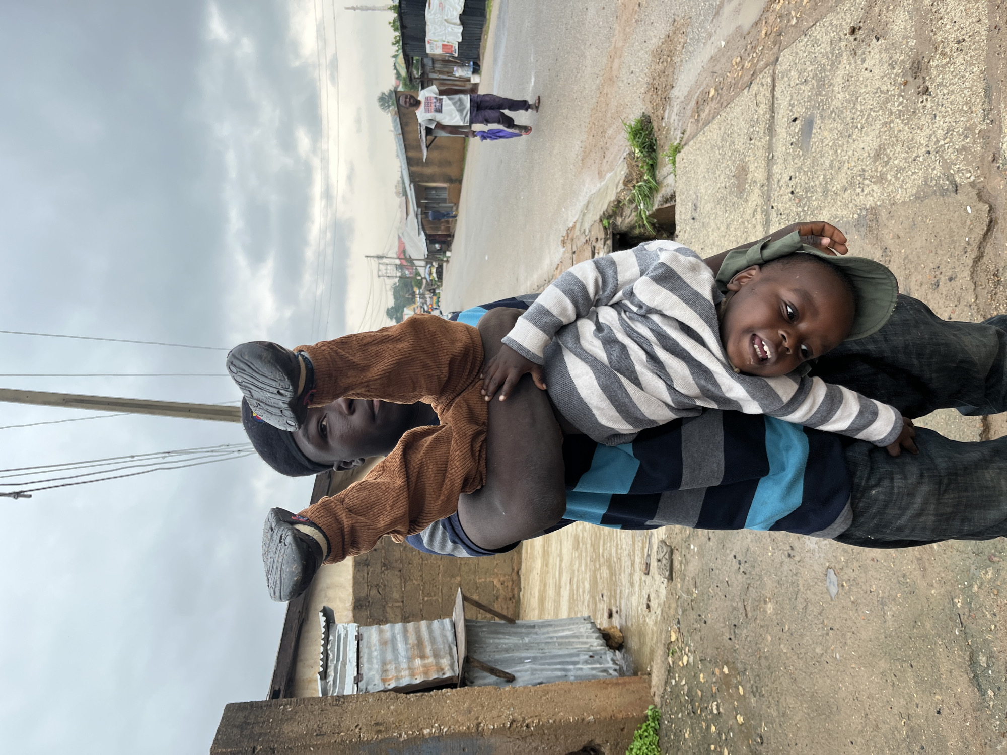This media file depicts an outdoor scene in Jos, Nigeria. The primary subjects are two individuals, an adult and a child. The adult is carrying the child, who is lying on their back, facing the camera. The child is wearing a striped long-sleeved shirt and a green hat, and appears to be smiling. The adult is wearing a striped shirt and dark pants, and their face is partially visible. They are walking on a paved area that appears to be a sidewalk or path next to a road.
In the background, a road stretches into the distance, lined with buildings that suggest a commercial or residential area. There are other people visible in the background, some walking along the road and others near the buildings. The scene is set during the day, with an overcast sky suggesting it might be cloudy or raining. The ground is wet in places, indicating recent rain. The overall atmosphere appears casual and everyday. There is no visible text in the image.