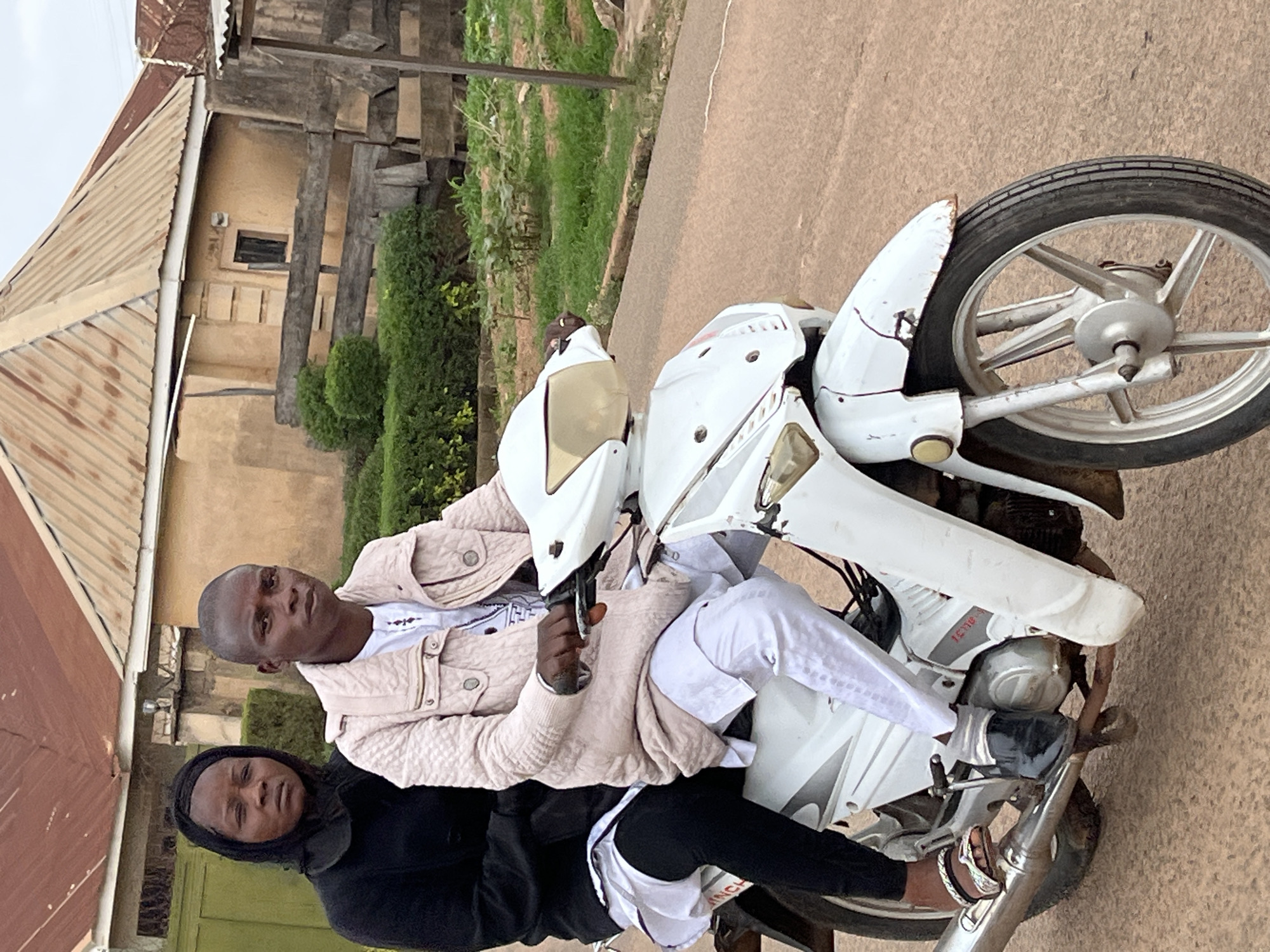 Two individuals, a man and a woman, are sitting on a white motorcycle on a paved road. The man is driving, wearing a light-colored jacket and a white shirt. The woman is sitting behind him, wearing a black hooded garment. The motorcycle is parked on a paved road adjacent to a building with a corrugated metal roof and a tan exterior. Green foliage is visible in the background. The location is identifiable as Jos, Nigeria, based on architectural styles and landscape features typical of the region.