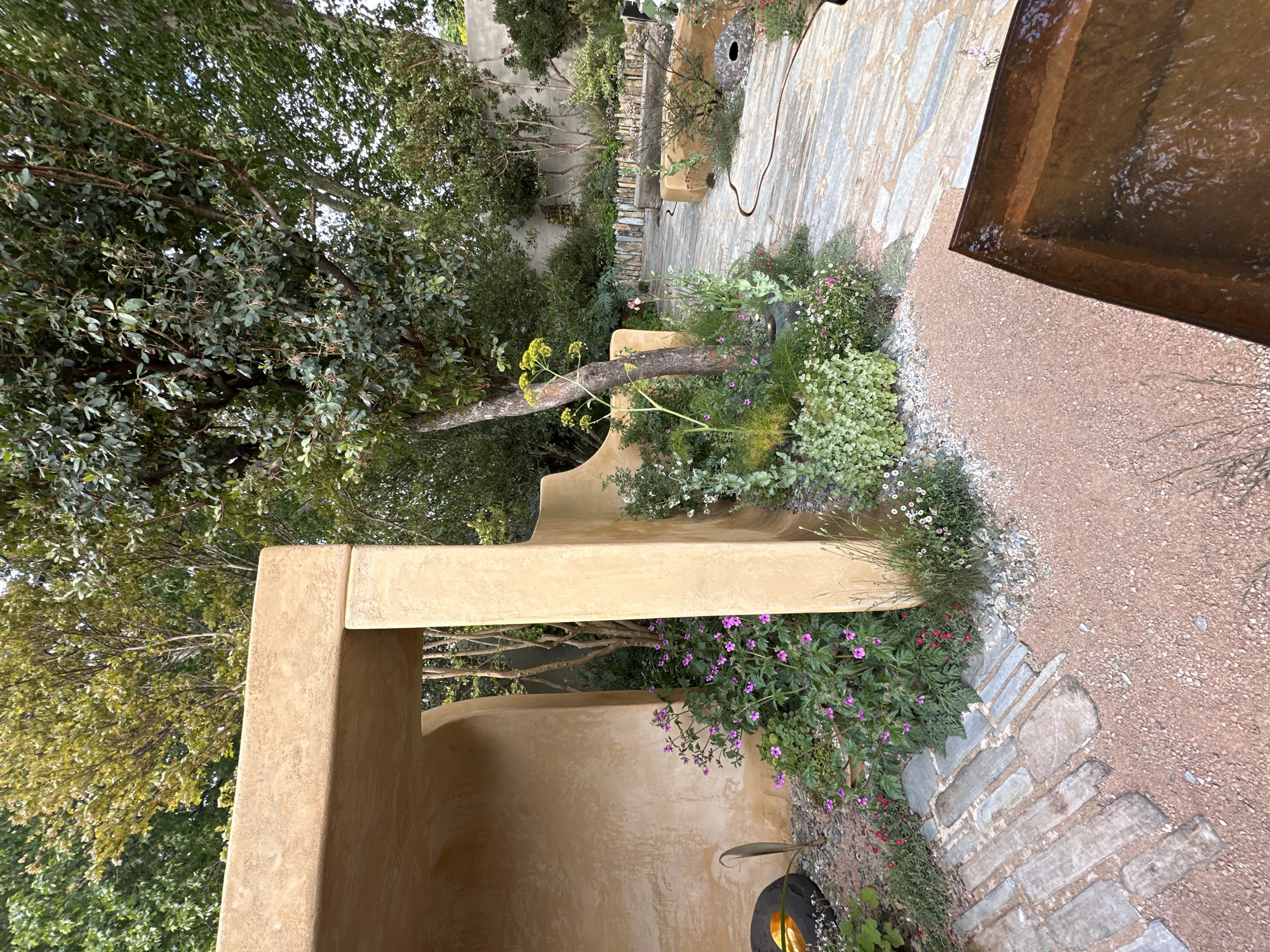 The image shows an outdoor garden space, possibly located in London, United Kingdom, given the style of the landscape design which often features in British gardens.
**Environment:** The garden has a Mediterranean-inspired aesthetic, with walls in a sun-baked earthen tone. A variety of plants thrive in the garden beds, including flowering perennials in purple, white, and red hues, interspersed with greenery. Trees provide shade, and the ground covering varies from cobblestone to gravel and stone slabs, creating a textured pathway.
**Objects:** A structure of light tan hue dominates the foreground. There appears to be a water feature in the upper right, with a metal frame. Plant containers are visible along the path.
**Subjects:** The main subjects are plants, garden architecture, and elements of the landscaping.
**Actions/Interactions:** None of the subjects are performing any actions. The plants and structures are arranged to create a visually appealing and functional outdoor space.
**People:** There are no people visible in the photograph.