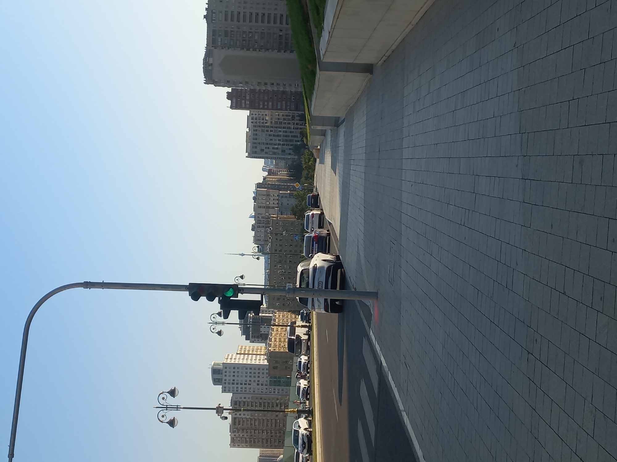 The image captures a street scene in Baku, Azerbaijan, with a clear blue sky overhead. In the foreground, a paved sidewalk runs alongside a road with several cars. A traffic light, displaying a green signal, is prominently visible on the right side of the road, mounted on a pole. The sidewalk is constructed with light gray square tiles. Further down the street, a pedestrian crossing is marked with white stripes. The background features a cityscape with numerous tall buildings, characteristic of urban development in Baku. Streetlights with decorative designs are also visible, adding to the urban ambiance. The overall impression is of a modern city on a bright, clear day.