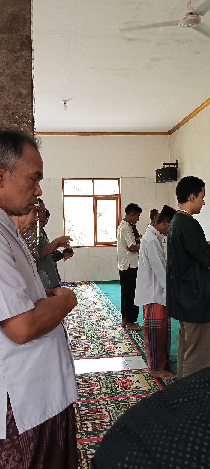 The image shows a group of men praying on prayer rugs in a mosque in Kecamatan Kutawaringin, Indonesia. The men are wearing traditional clothing and are standing in a row facing the direction of prayer. The walls are white and there is a window with a view of the outdoors. The ceiling is white and there is a fan hanging from the ceiling. The floor is tiled and covered with prayer rugs. The mosque is a simple, but well-maintained building. There is a speaker attached to the wall. A man in a light blue shirt is standing in the foreground, his hands are crossed in front of him. He is looking to his left. There is a man in a gray shirt in the background. He is looking towards the camera and he is holding his hands together in front of him. The scene is peaceful and serene.