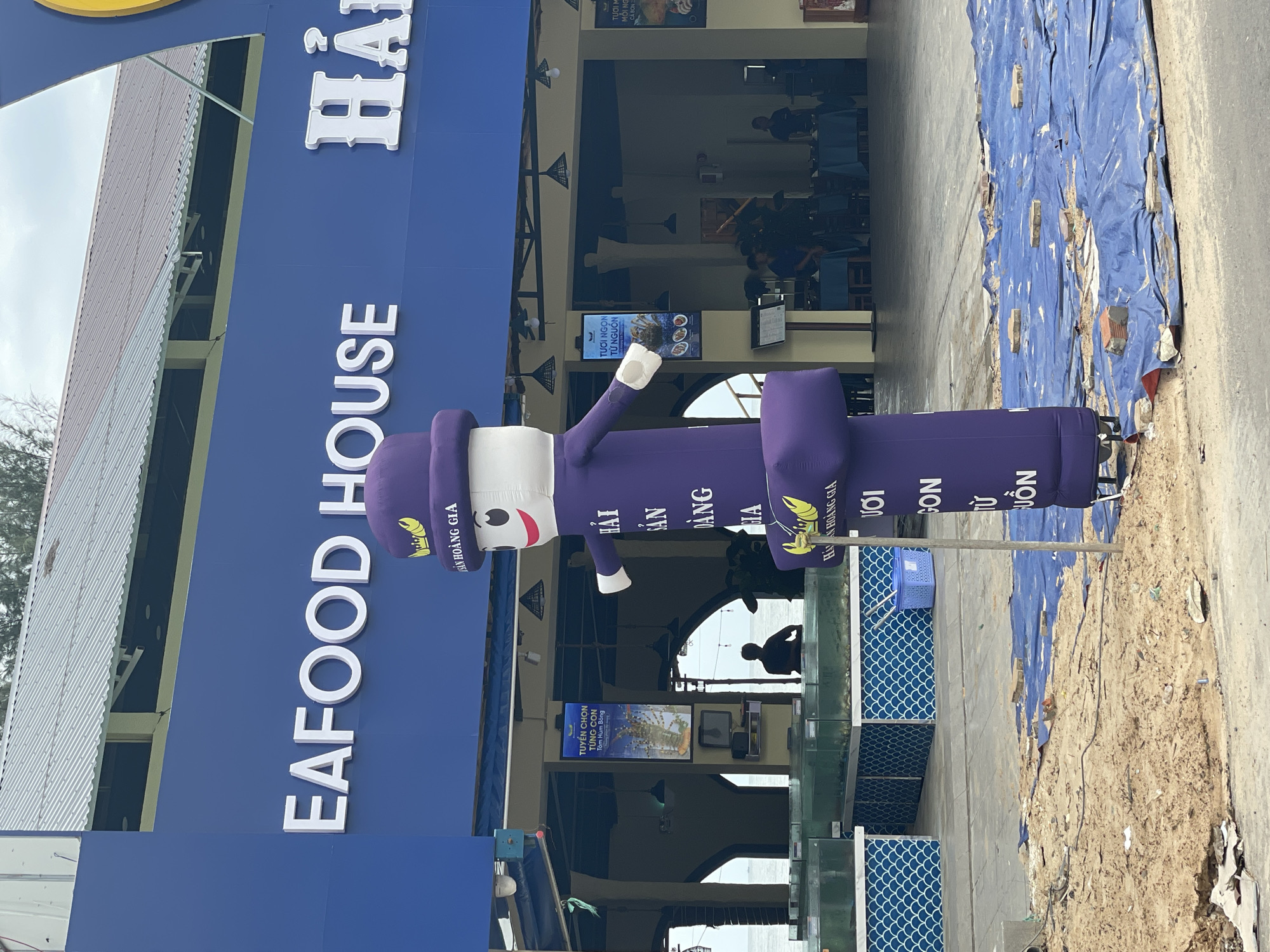 Here is a detailed description of the image:
**Scene:** The image shows the exterior of a restaurant named "Hải Sản Hoàng Gia Seafood House" in Phu Thuy Ward, Vietnam. The restaurant's name is prominently displayed on a blue wall.
**Objects:**
* **Advertising inflatable:** A tall, purple inflatable tube man with a cartoon face and the restaurant's name is standing outside, likely attracting customers.
* **Restaurant facade:** The building is painted blue and features white lettering for the restaurant's name.
* **Aquariums:** There are multiple glass aquariums displayed on the outside of the building.
* **Promotional material:** A digital screen displays advertisements for the restaurant, including images of seafood.
**People:**
* Several people are inside the restaurant.
* One person is silhouetted at the back of the restaurant.
**Environment:** The image is taken outdoors, with a street or sidewalk visible. The sky is cloudy.