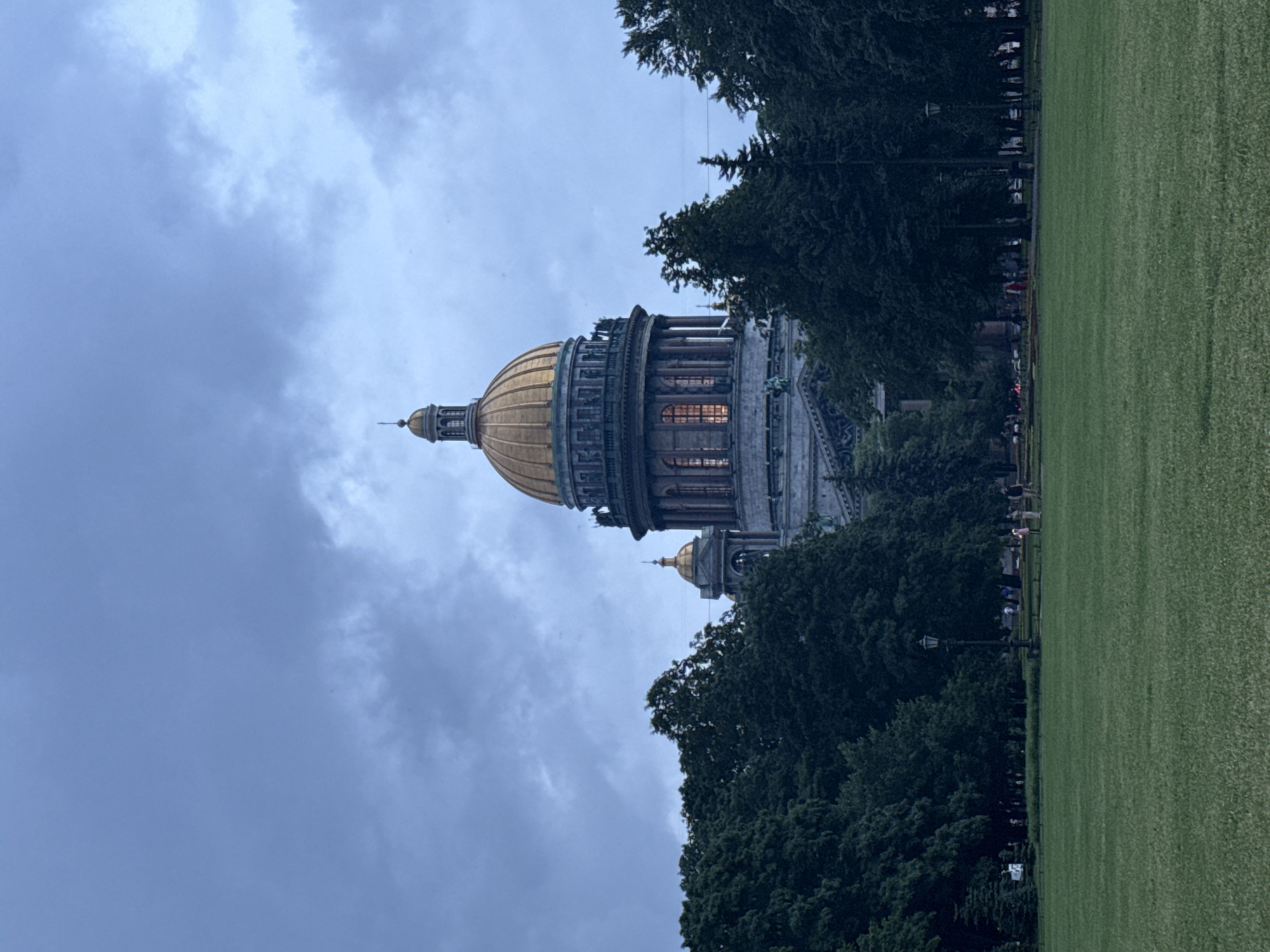 The image captures Saint Isaac's Cathedral in Saint Petersburg, Russia, under a dramatic, cloudy sky. The cathedral, a prominent landmark, features a large, gilded dome that catches the subdued light. Dark green trees partially obscure the lower sections of the building, with their dense foliage forming a natural border. In the foreground, a vast, well-maintained lawn stretches out, appearing lush and green. Several figures are visible on the lawn and near the trees, some walking and others standing, suggesting a public park setting. A lamppost stands on the edge of the lawn, adding a touch of urban infrastructure to the scene. The overall atmosphere is serene, with the subdued lighting and cloudy sky contributing to a sense of calm and grandeur.