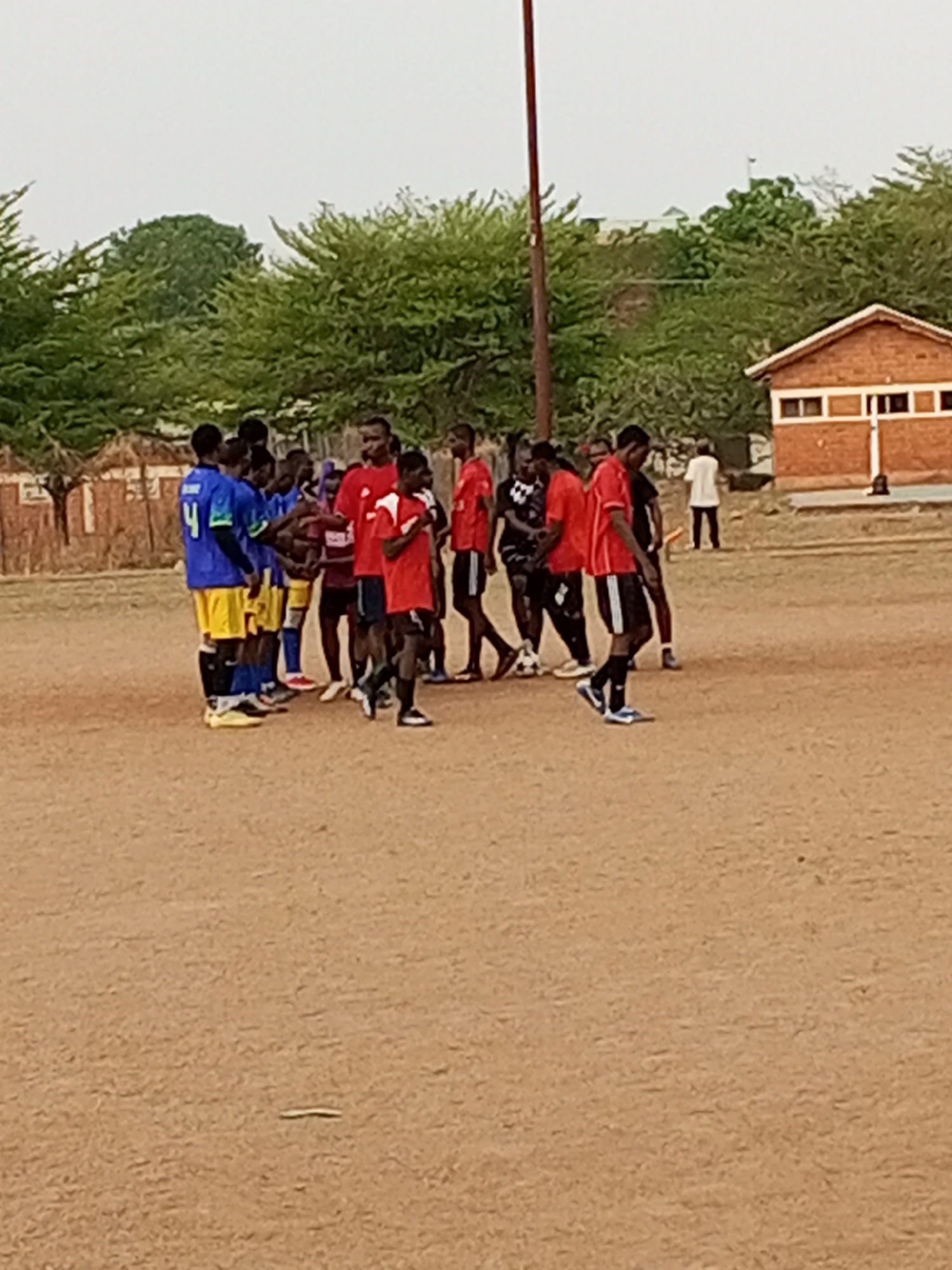 The image shows a group of soccer players, some in blue and yellow uniforms and others in red and black uniforms, standing on a dirt field. There is a small brick building in the background to the right of the soccer players and a tall pole on the left side. The setting appears to be outdoors, with trees and other vegetation visible in the background. Based on the context of previous interactions, the location is potentially in the city of Minna, Nigeria. The teams appear to have just finished a game and are interacting with each other.