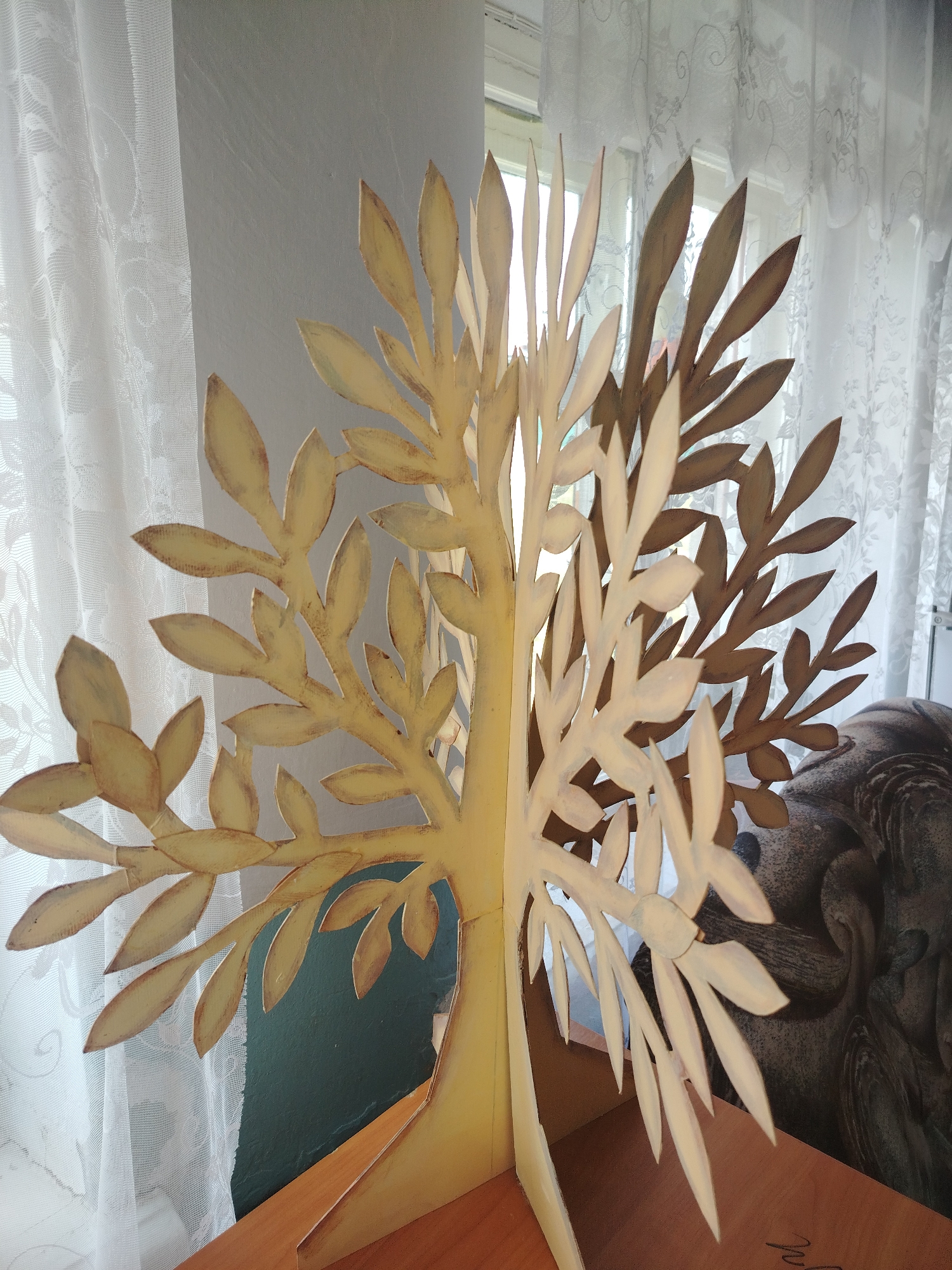 The image depicts a freestanding, two-dimensional, stylized tree sculpture with light yellow and brown leaf-like cutouts. The sculpture is positioned on a wooden surface, likely a table or shelf, in front of a window with sheer white lace curtains. The background shows a muted green wall and a portion of what appears to be a stone or carved object, possibly a bust or statue, to the right of the tree. The scene is indoors, suggesting it is a decorative piece within a room. There are no people visible in the image. The overall lighting suggests an indoor setting with natural light coming from the window. The location of Klyuchi, Russia, is not visually discernible from the provided image.