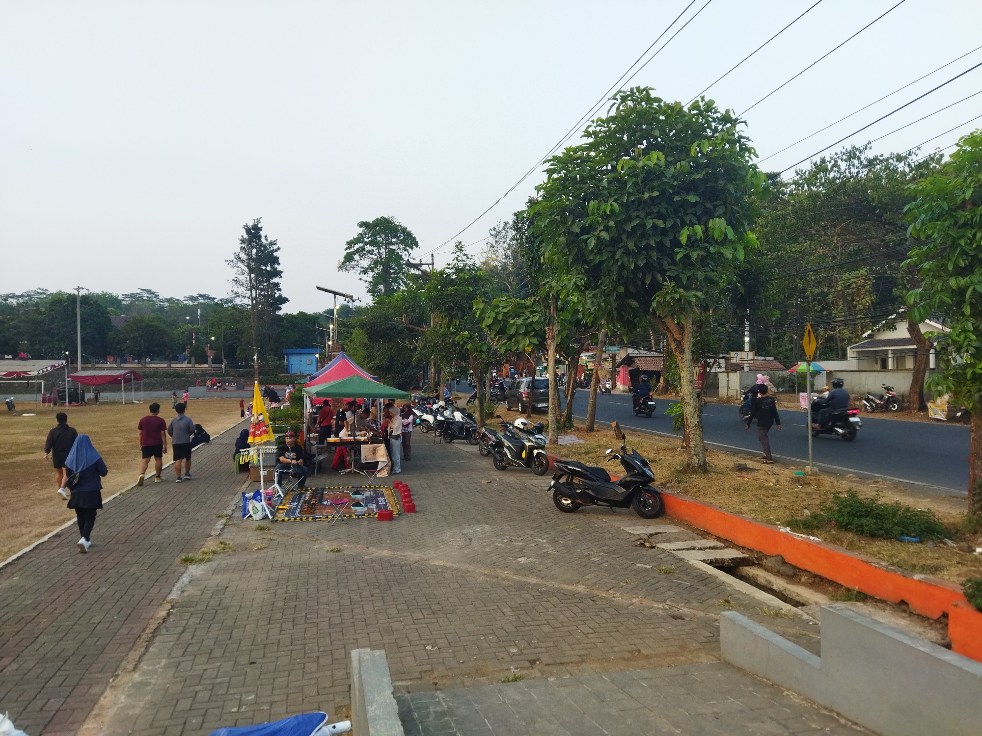 The image depicts a street scene in Semarang, Indonesia, with a paved pedestrian walkway bordering a street. Several people are visible, some walking along the path and others gathered near market stalls. Motorcycles are parked along the street and are also being ridden by some individuals. The environment is characterized by trees and some buildings in the background. The sky is overcast, suggesting it might be dawn or dusk. The market stalls appear to be selling food or other goods. The overall atmosphere suggests a busy, public area with both pedestrian and vehicle traffic.