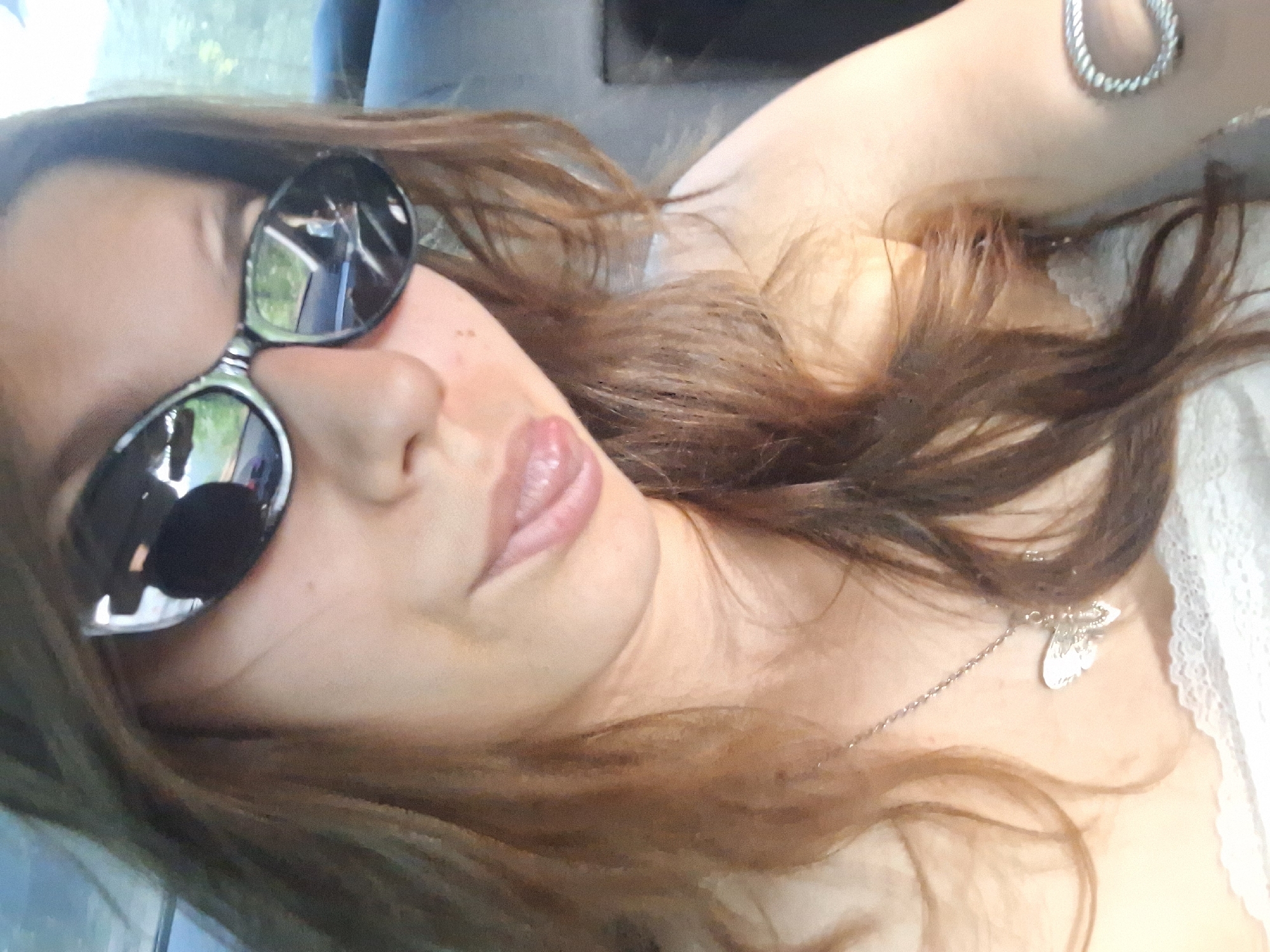 A young woman is taking a selfie in a car. She is wearing black sunglasses and has her tongue sticking out. She has long brown hair and is wearing a necklace with a butterfly pendant. The car appears to be in motion, as evidenced by the reflection in her sunglasses, which shows trees and a road. The image is taken from a slightly high angle, looking down at the woman. There is no visible indication of the city of Koprivshtitsa, Bulgaria.