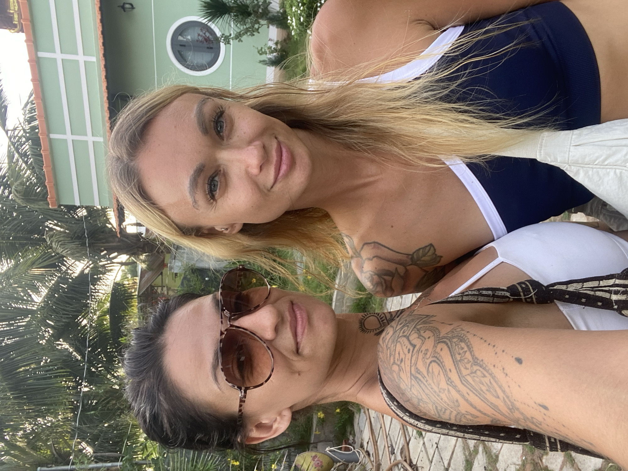 Two women are posing for a selfie outdoors. The woman on the left has dark hair pulled back and is wearing large, leopard-print sunglasses. She is also wearing a white top and has extensive tattoos on her arm and chest, including a sun design on her neck. The woman on the right has long blonde hair and is wearing a dark blue and white athletic top. She is smiling broadly. Behind them, a light green building with white trim and an orange-tiled roof is visible, along with palm trees and tropical foliage. The ground appears to be paved with stone. The scene is consistent with the location of Mũi Né, Vietnam, known for its tropical climate and beachside resorts.