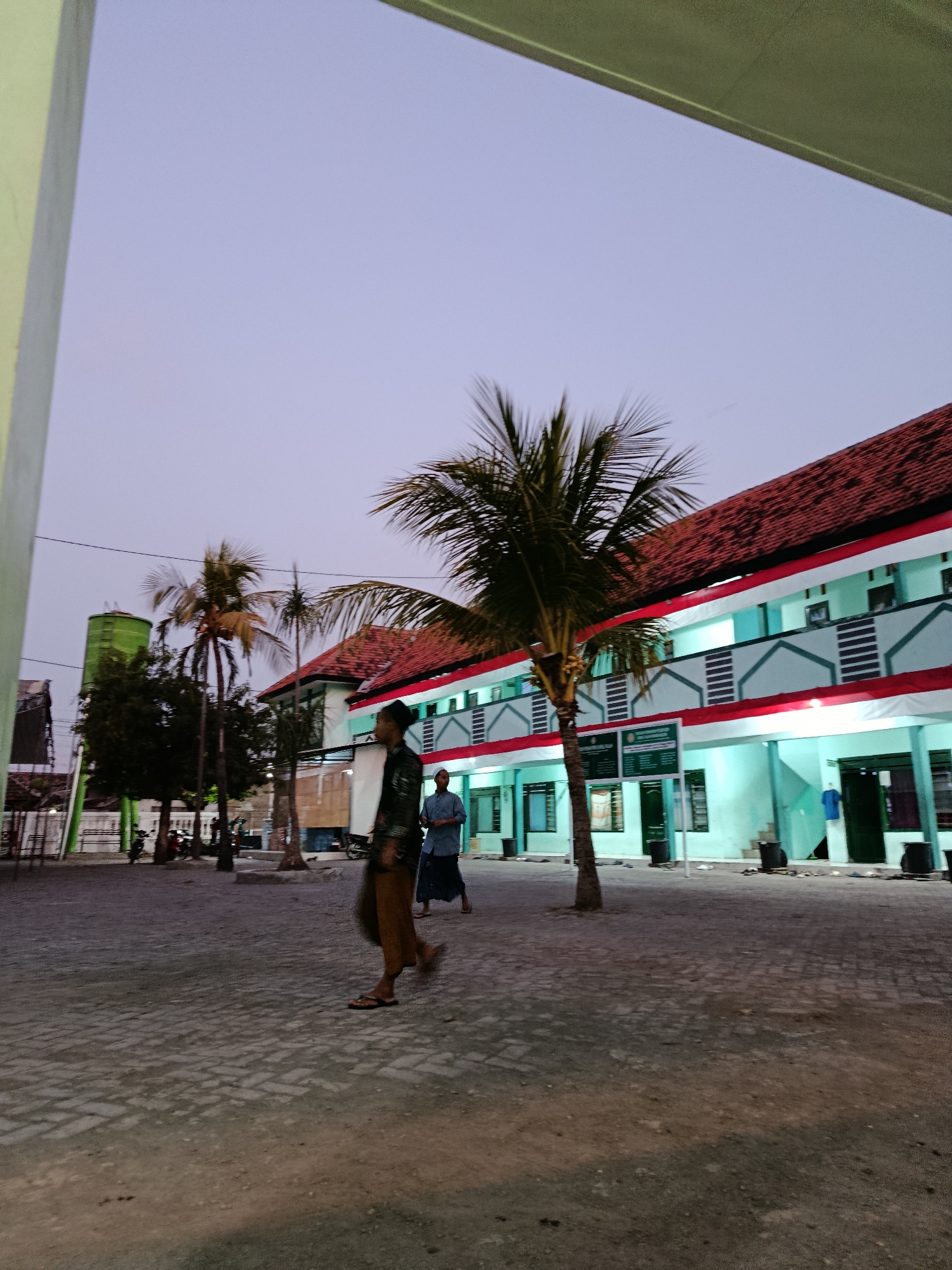 This is a medium shot, low-angle photograph taken at dusk in Ramban Wetan, Indonesia, depicting the exterior of a building that appears to be a school or religious institution. The building is multi-storied with a light blue facade, white trim, and a red tiled roof. Several windows are visible on each floor, some with dark shutters. A prominent palm tree stands in the foreground, its fronds reaching towards the twilight sky. Two men are walking in the courtyard, one in the foreground wearing a brown sarong and a dark patterned shirt, the other further back in a light blue shirt and dark pants. The courtyard is paved with interlocking grey bricks. In the background, a tall green cylindrical water tower is visible, along with other trees and structures. The overall atmosphere is calm and serene, indicative of a peaceful evening.