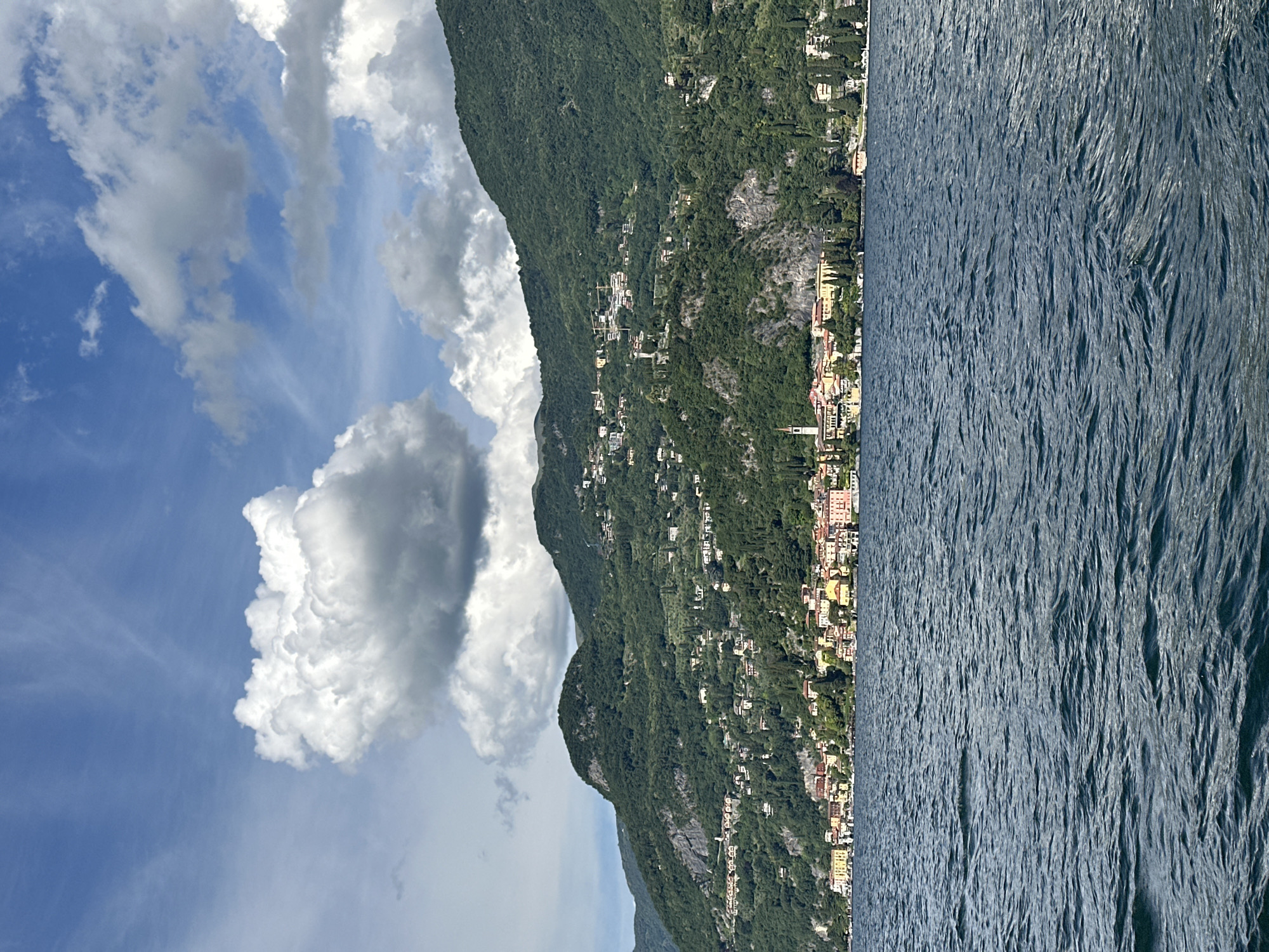 The image displays a scenic landscape depicting a body of water, a town, and mountainous terrain under a partly cloudy sky, characteristic of Gittana, Italy. The upper portion features a bright blue sky partially obscured by large, billowy white cumulus clouds, with some darker, shadowed clouds present towards the upper right. The middle ground is dominated by lush green mountains densely covered in trees and vegetation. Numerous residential structures, characterized by various colors and terracotta roofs, are scattered across the mountainsides, ascending from the shoreline. A distinct town or village extends along the base of the mountains at the water's edge, featuring closely packed buildings with colorful facades (yellows, oranges, and pastels) and a prominent, slender church tower or campanile on the left side of the town. The lower portion of the image shows the surface of a deep blue lake with visible ripples and small waves, suggesting slight movement or wind on the water. No individuals, specific actions, or interactions are discernible.