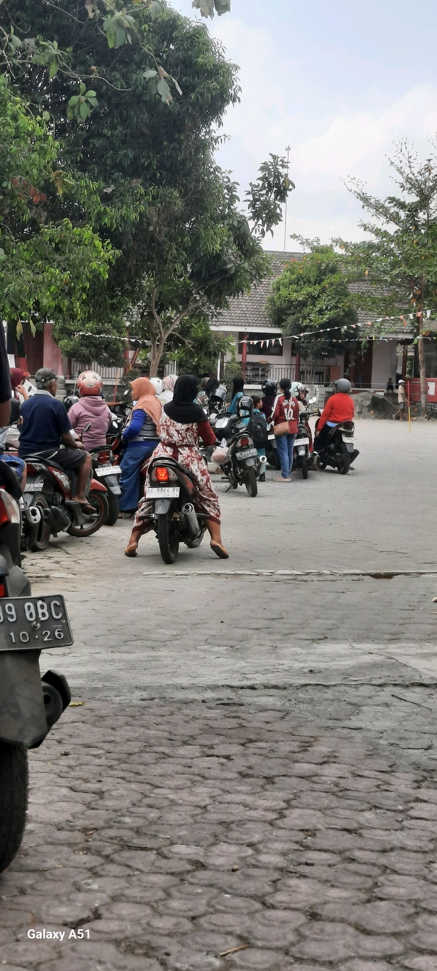 In the city of Tangkil, Indonesia, a group of people are gathered on motorcycles in what appears to be a parking area. The scene is set outdoors with trees and buildings in the background, suggesting it could be near a school or community center, possibly indicated by the small flags strung across the buildings. The pavement is a mix of asphalt and interlocking stone blocks. The sky is overcast, hinting at a cloudy day. The individuals, both male and female, are mostly dressed in casual clothing and many are wearing hijabs, which is common in Indonesia. Some are seated on their motorcycles, while others appear to be waiting or about to depart. The motorcycles are parked closely together, creating a sense of a busy gathering. The overall atmosphere is one of everyday activity, likely a routine departure or arrival.