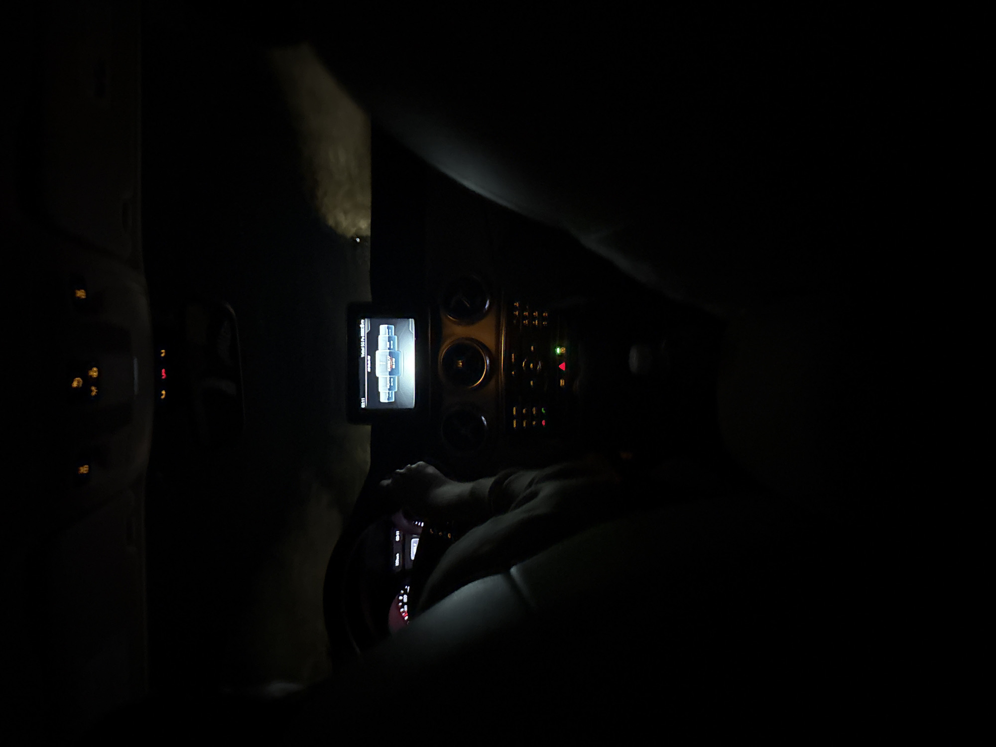 The image depicts the interior of a car at night. The dashboard and steering wheel are visible. A person is behind the wheel. A screen displaying an image is mounted on the dashboard. The location is unknown.