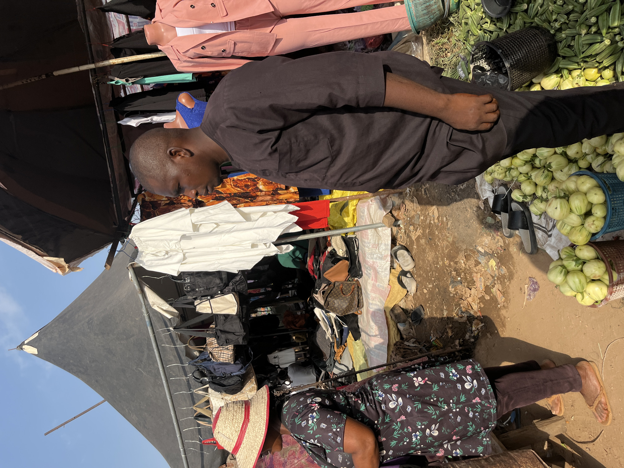 The image depicts a street market scene in Nyanya, Nigeria. Two individuals are visible. In the foreground, a person wearing a dark floral-patterned dress and brown trousers is sitting with their legs extended. They are also wearing a straw hat with red trim. Behind them, and to their left, a person wearing a dark brown tunic and black trousers is leaning forward, appearing to be looking at something below.
The scene is cluttered with various objects. In the background, racks of clothing, including pink jackets and pants, are displayed. Bags and other merchandise are also visible, some hanging from structures and others piled on the ground. Baskets filled with produce, likely okra and green, bulbous vegetables, are prominent. The ground is a mix of dirt and scattered debris. A black canopy tent provides shade.
The activity appears to be the daily operation of a market. The time of day seems to be during daylight hours, as indicated by the strong sunlight casting shadows. The weather appears to be warm and sunny. The overall impression is one of a busy, informal marketplace. No visible text is discernible in the image.