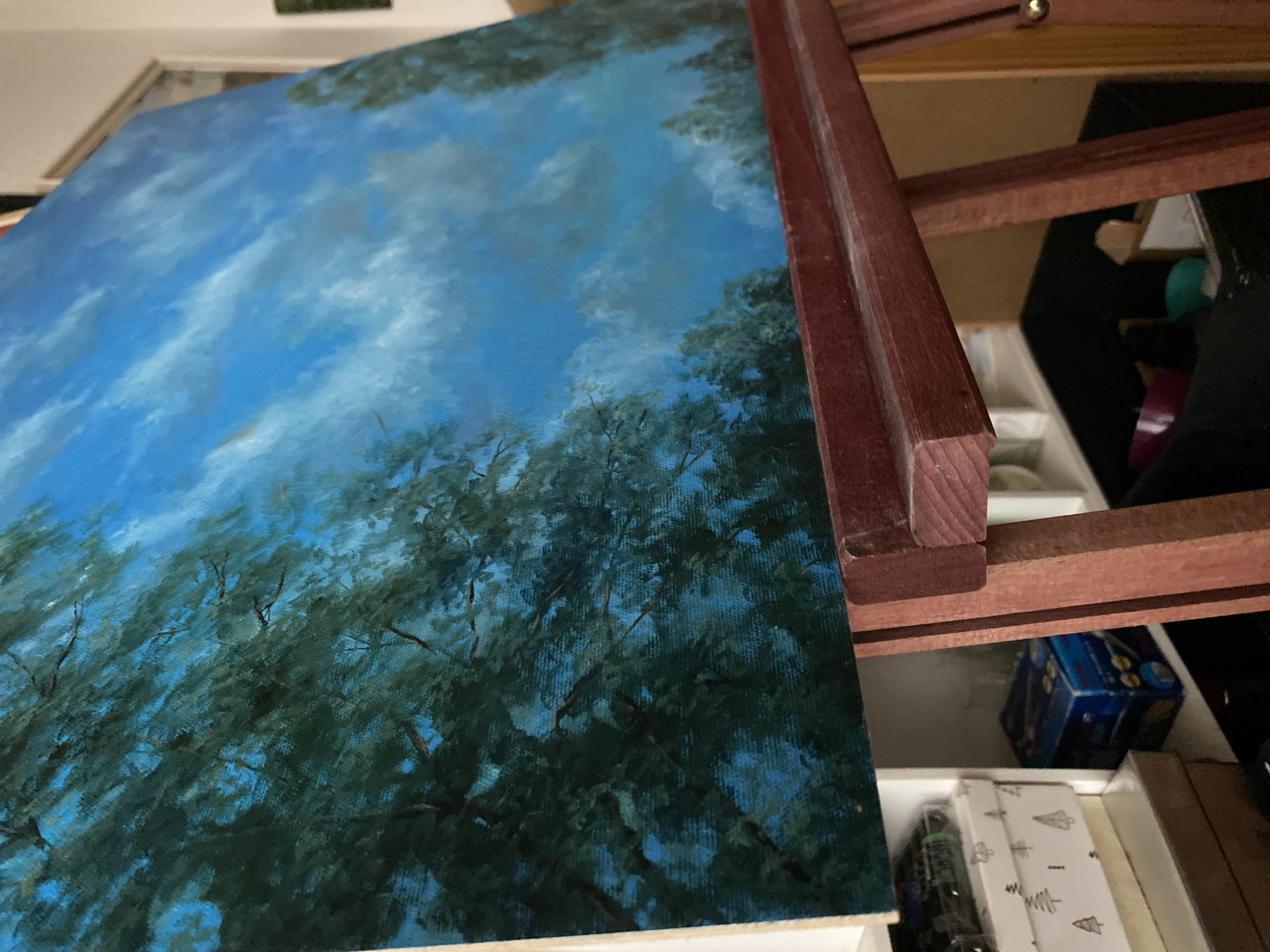 An oil painting of trees and a blue sky with clouds is displayed on a wooden easel. The painting is angled to the right, with dark green trees filling the left side of the canvas and a bright blue sky with white clouds occupying the right. The easel is made of dark red wood and is positioned in front of shelves that hold various boxes and containers. The background appears to be an indoor setting, possibly a room in Bílina, Czechia, with a hint of a wall and a framed picture visible on the right. The lighting suggests an indoor environment, likely natural light coming from a window.