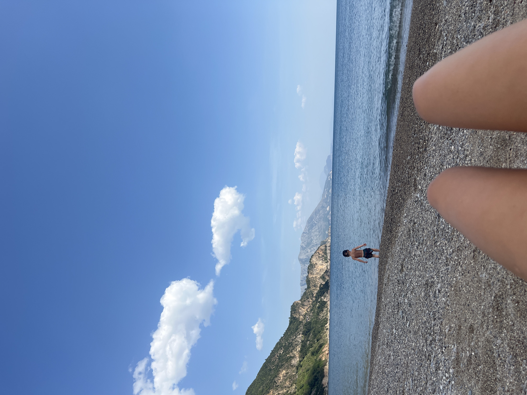 The image displays a sunny day at a pebbly beach or lakeside in Prijevor, Montenegro. The horizon is visible, separating a vast, clear blue sky with scattered white cumulus clouds from a calm body of water. Distant mountains, covered in green vegetation and rock, frame the background, rising directly from the water's edge on the left.
In the mid-ground, a young male individual, appearing to be a child or young adult, stands in the shallow, clear water near the shore. He is wearing dark swim shorts and dark sunglasses perched on his head. He faces generally away from the camera, towards the open water. The foreground prominently features the knees and lower thighs of a person, presumably the photographer, suggesting a first-person perspective from someone relaxing on the beach.
The shore consists of dark, smooth pebbles. The water near the edge is calm, with minimal wave activity. The overall impression is one of a peaceful and idyllic summer day, with bright natural light illuminating the scene. There is no visible text in the image.
