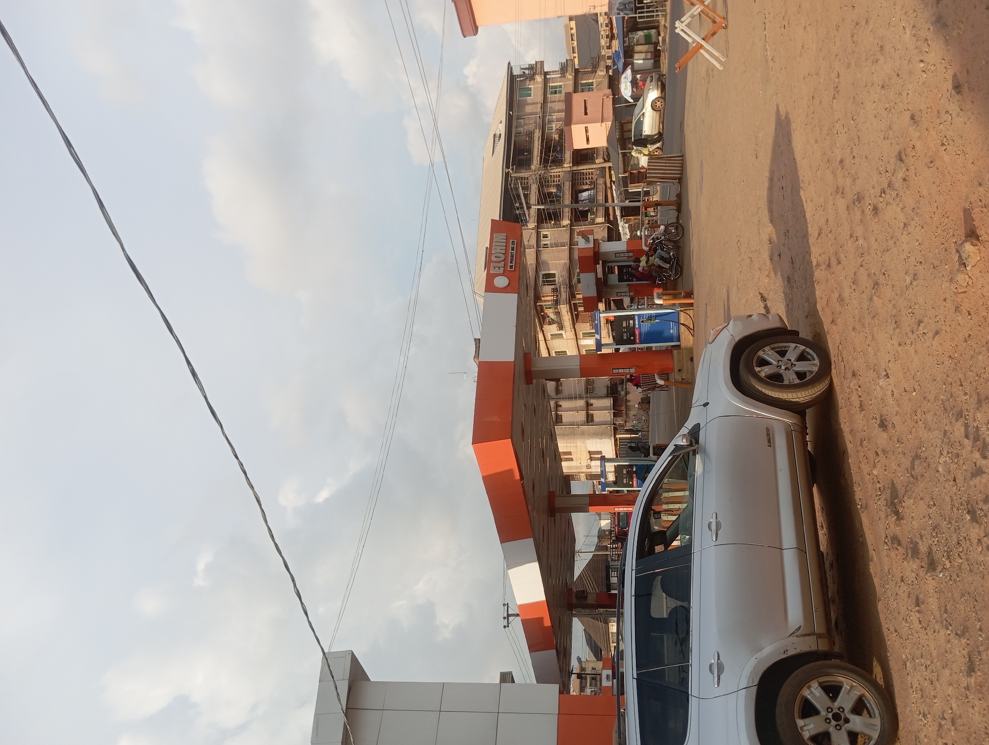 The image depicts a scene in Nkpor, Nigeria, showing an Elokim Oil filling station. A silver SUV is parked in front of the station. Adjacent to the filling station is a building with multiple floors and exposed balconies, suggesting residential or commercial units. Motorbikes and people are visible near the filling station, and various goods or materials are arranged in front of the building next door. The road surface is dusty, and utility lines traverse the sky. The sky shows partly cloudy conditions.