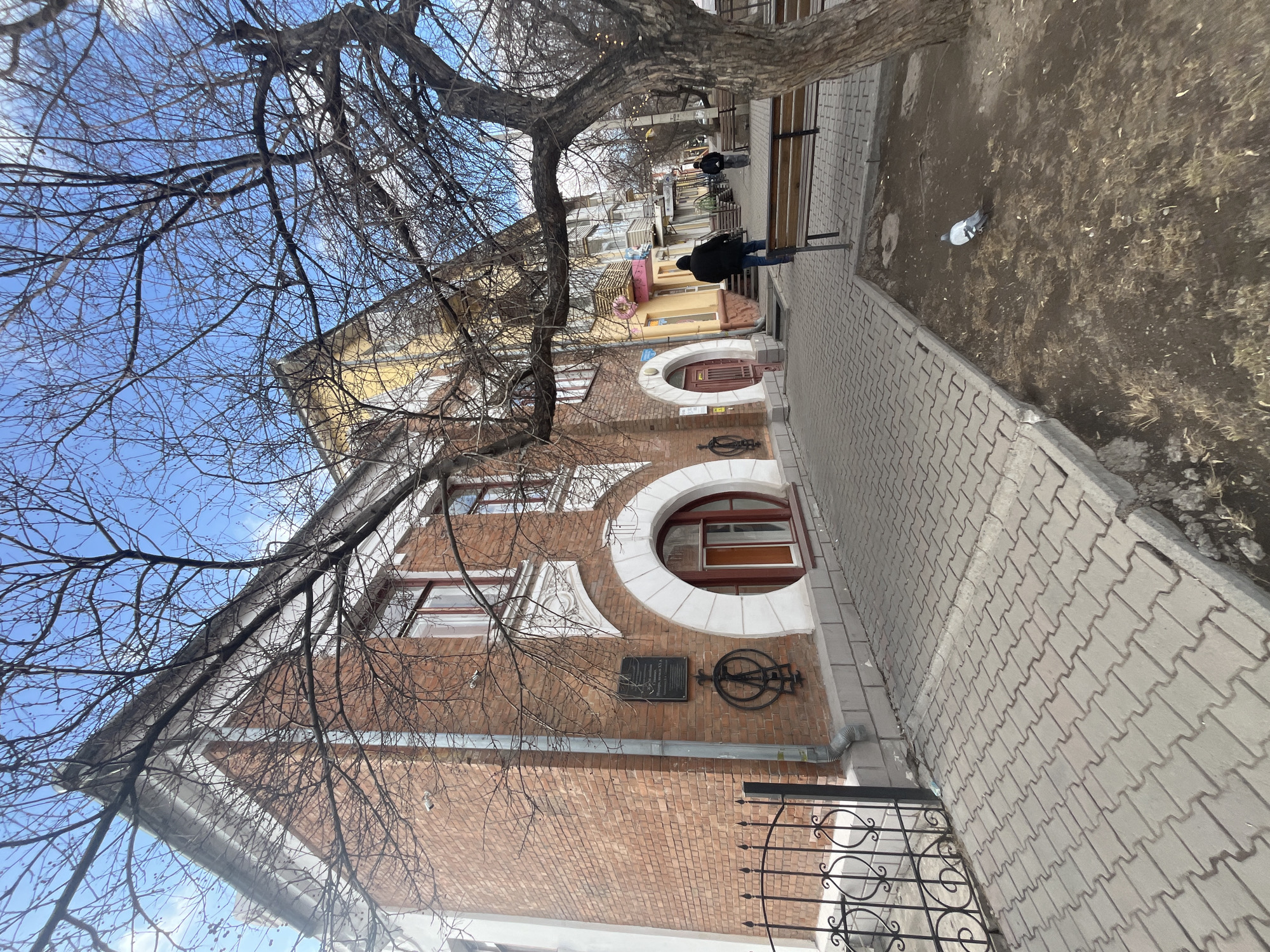 The image shows a street scene with a brick building on the left side, featuring distinctive round windows with white trim. A plaque and a decorative metal emblem are mounted on the building's exterior. A sidewalk made of paving stones runs along the side of the building, and a wrought-iron fence is partially visible near the bottom right corner. A tree with bare branches dominates the upper left portion of the frame, obscuring parts of the sky and other buildings in the background. Further down the sidewalk, a few individuals are walking, and benches are placed along the path. The street appears to be in Unknown, Unknown.