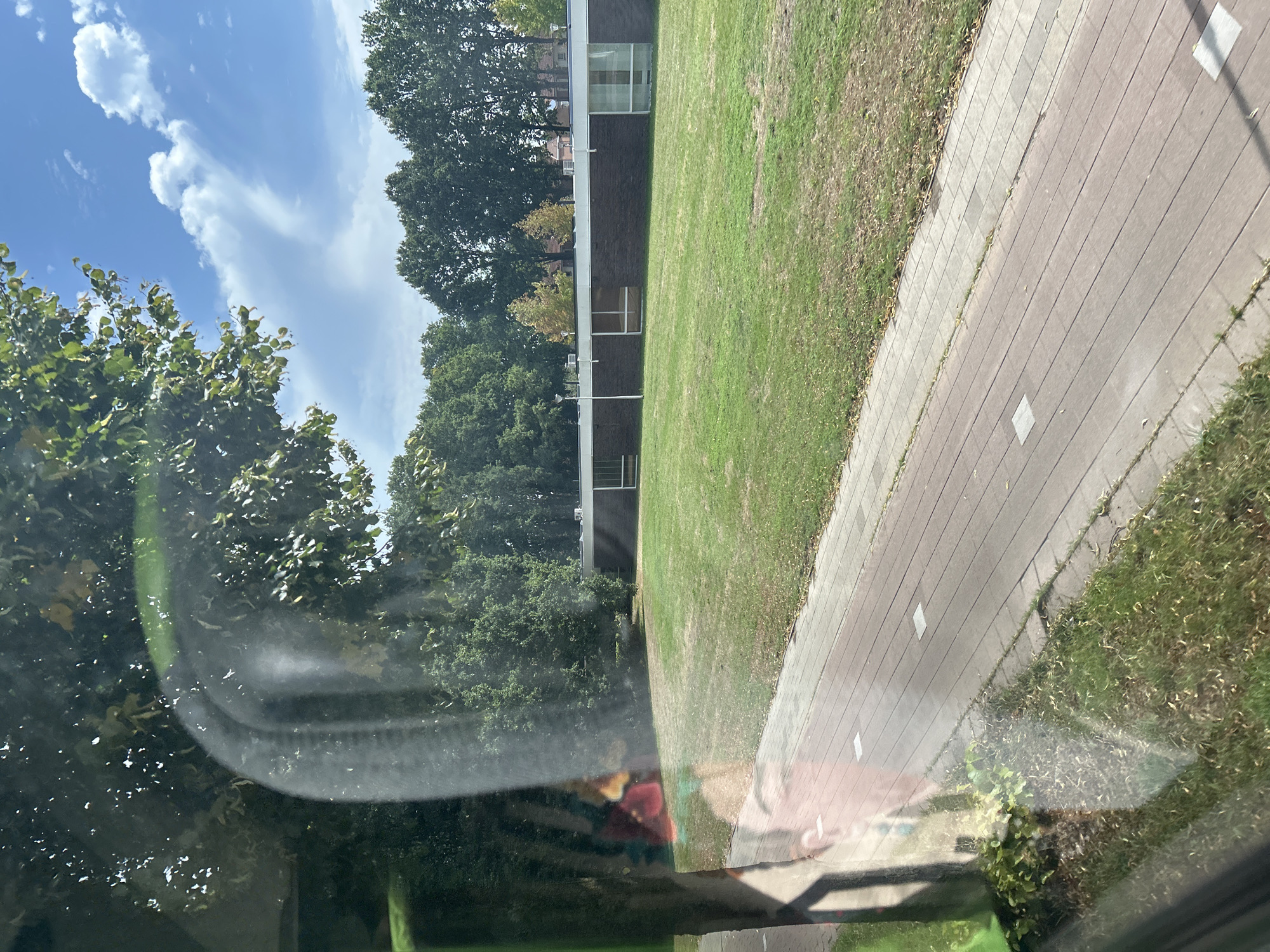 The image captures a reflection on a window, showing a suburban scene in Tilburg, Netherlands. A brick building with large windows and a white roofline is visible in the background, surrounded by lush green grass. A paved path runs in front of the building, bordered by grass. Trees with dense green foliage line the horizon, with some autumnal yellow leaves visible on a few trees. The sky is a clear blue, dotted with white clouds. In the foreground, the reflection shows a tree trunk and a hint of foliage. There are no people or animals clearly visible in the scene, though a faint reflection of a person's face is present on the left side of the window.