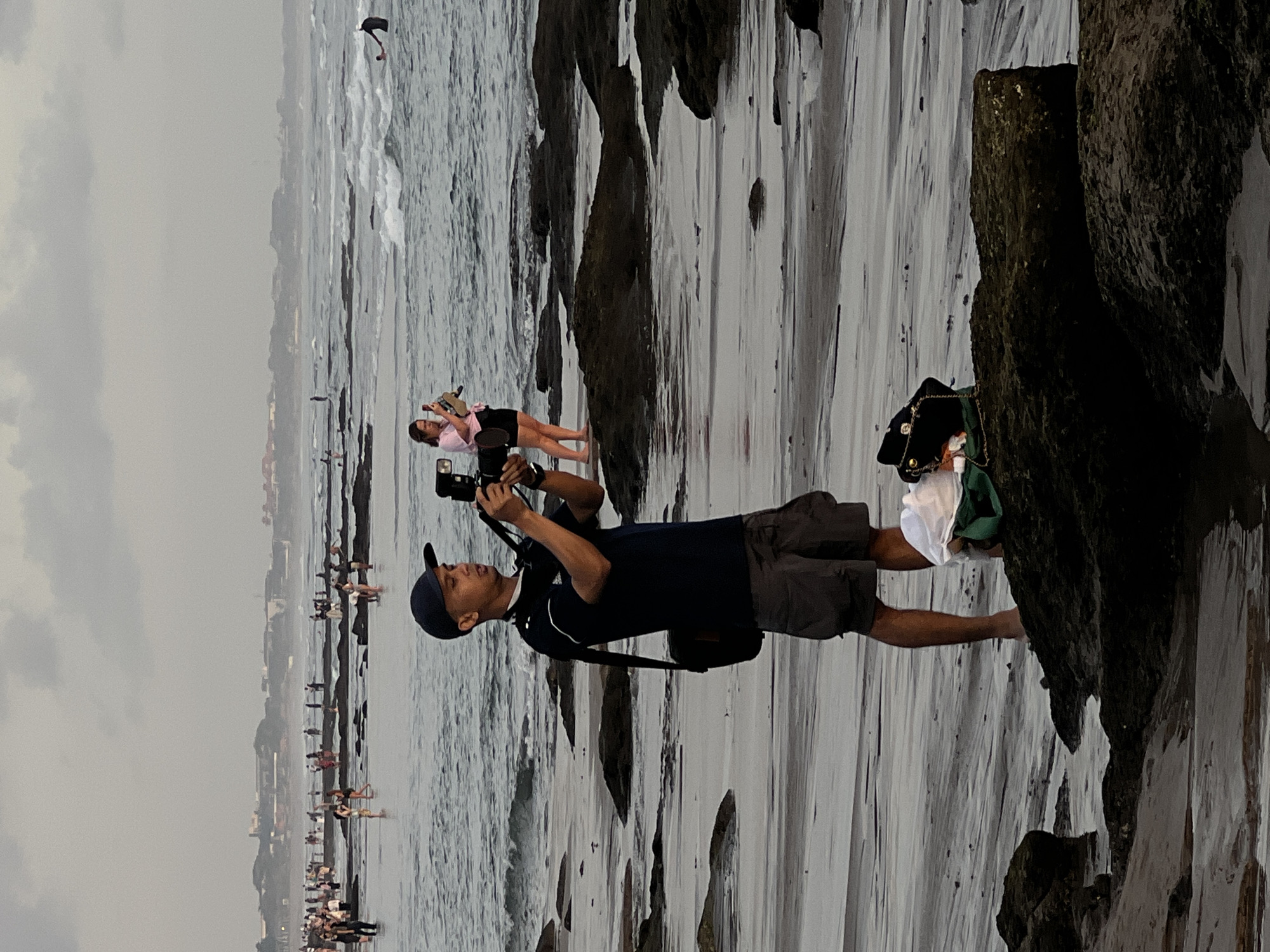 A man is taking photographs on the beach at Canggu, Indonesia. The man is standing barefoot, wearing a dark blue shirt, gray shorts, and a baseball cap. He holds a camera to his eye. In the background are other people at the shore, and rock formations along the beach. Further in the background are buildings along the beach on the shore. The sky is overcast.