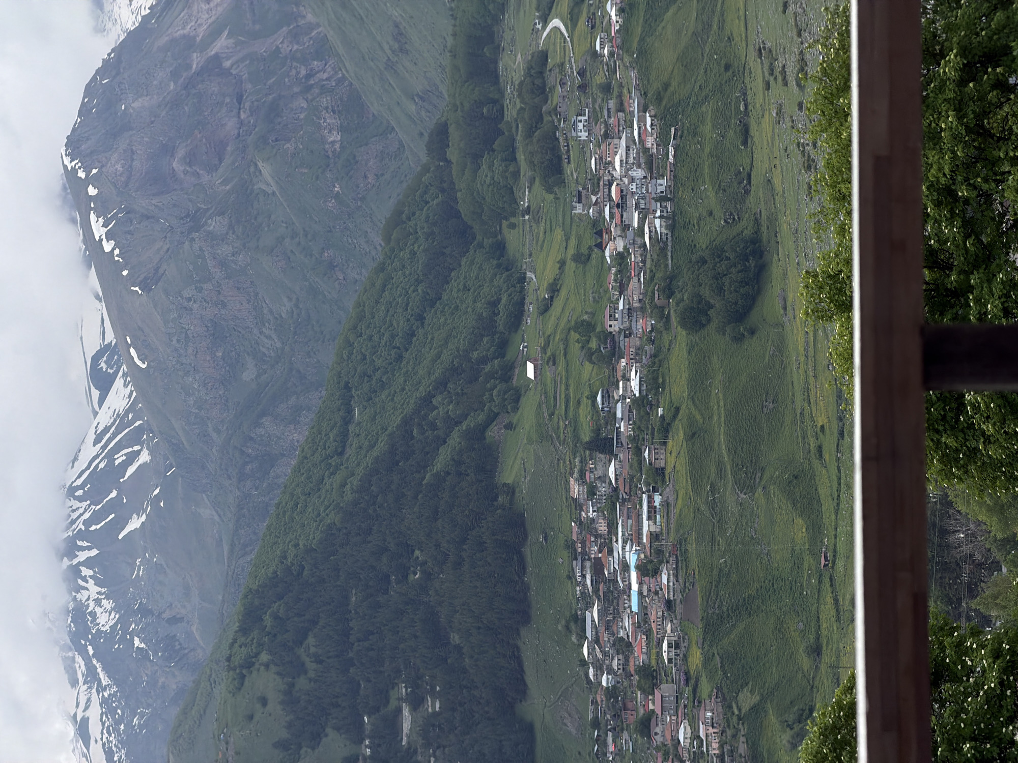 This media file depicts a high-angle view of a village nestled in a mountainous landscape. The scene is dominated by rolling green hills and dramatic, rocky mountains in the background, some of which are dusted with snow. A dense cluster of houses with red and white roofs forms the village, spread out along the contours of the terrain. A winding road is visible, snaking through the village and the surrounding landscape. The overall setting suggests a rural, mountainous area, likely in a temperate climate given the lush greenery.
The time of day appears to be daytime, with soft lighting suggesting either early morning or late afternoon. The weather seems overcast or hazy, as indicated by the diffused light and the presence of clouds or mist around the snow-capped peaks. There are no visible people or explicit activities taking place, creating a sense of stillness and tranquility. The presence of trees and vegetation suggests a natural, possibly remote location. The image also includes a wooden structure in the foreground, possibly a balcony or fence, which frames the view and adds a sense of perspective. No text is visible in the media file.