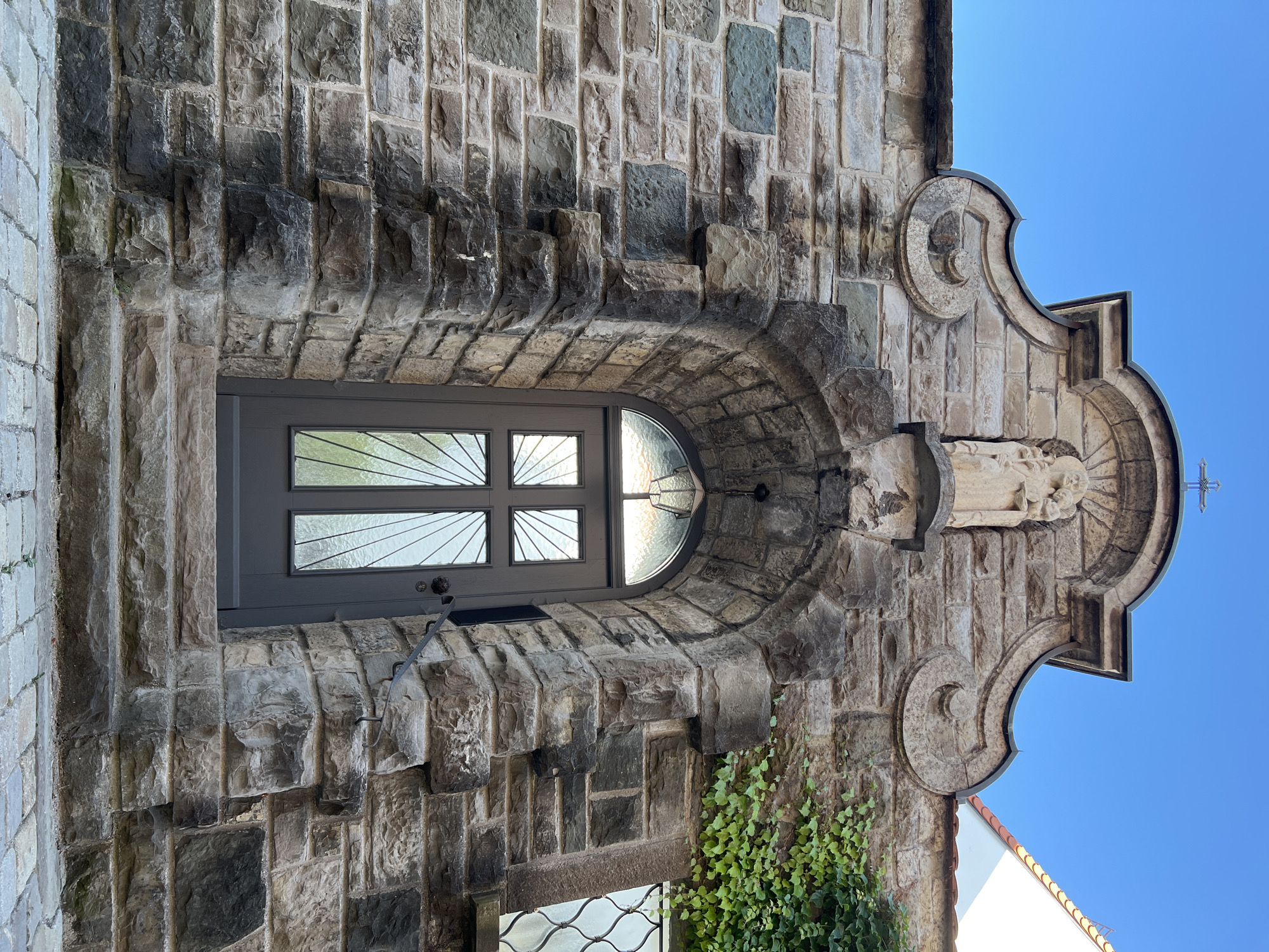An eye-level, medium shot captures the ornate stone facade of a building in Immenstadt im Allgäu, Germany, focusing on a Gothic-style arched doorway. The wall is constructed of rough-hewn, varied-colored stones, with the doorway framed by larger, darker stones that create a stepped effect. Above the arch, a stone sculpture depicts a religious figure, possibly the Virgin Mary, holding a child, and a cross tops the structure. The door itself is dark gray with a semi-circular glass top featuring a radiating pattern. The surrounding environment is clear, with a bright blue sky above and a glimpse of green ivy on the right side of the building. Cobblestone paving is visible at the base of the steps leading to the door.