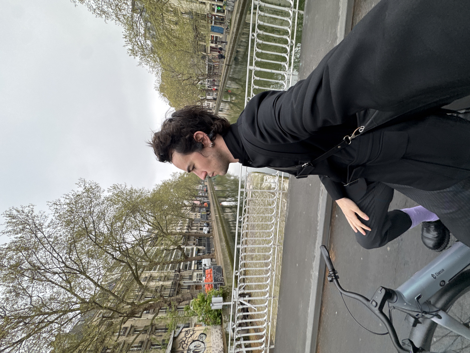 A person stands on a bridge in Paris, France, with a bike. The person, facing left, is wearing a black outfit, including pants, shoes, jacket, and a purple sock. The bike is light gray. The bridge has a white railing, and a canal runs below, lined with buildings and trees. The sky is overcast. Graffiti is visible on the structure of the bridge.