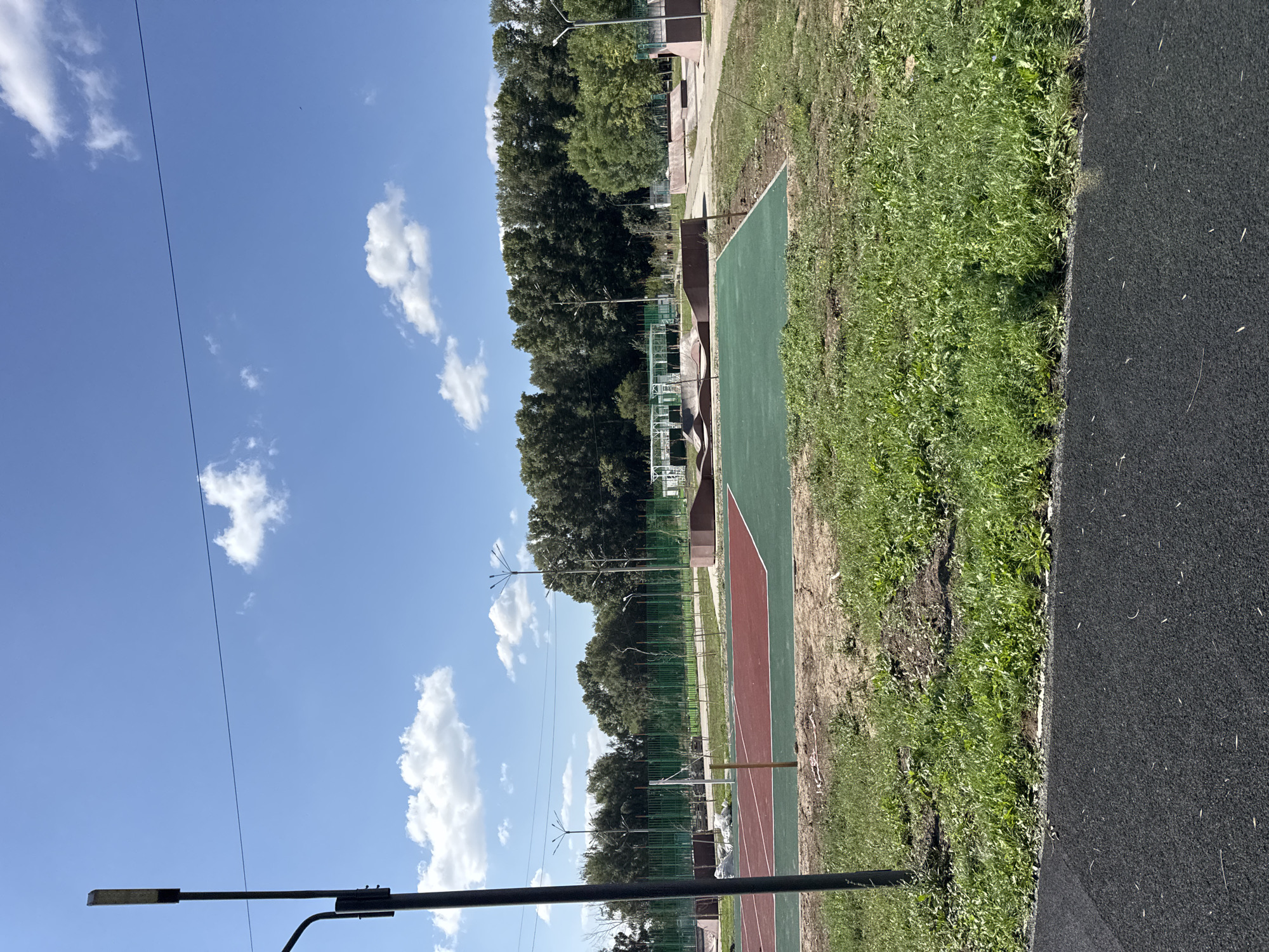 This is an outdoor photograph taken on a sunny day, with a bright blue sky dotted with white clouds. The image captures a newly developed sports complex in the city of Oskemen, Kazakhstan. In the foreground, a black rubberized track runs horizontally across the bottom of the frame. Beyond the track, a sports court with red and green playing surfaces is visible, divided by a thin white line. A volleyball net is set up on the court. A tall, dark lamppost stands to the left of the court.
In the middle ground, a skate park with concrete ramps and structures is laid out. A green fence encloses a portion of the sports complex. Behind the skate park and fence, a dense line of green trees forms the backdrop, suggesting a park or forested area. The ground in and around the sports areas appears to be a mix of sand and grass. Several other lampposts are sparsely distributed throughout the complex. There are no people visible in the image.