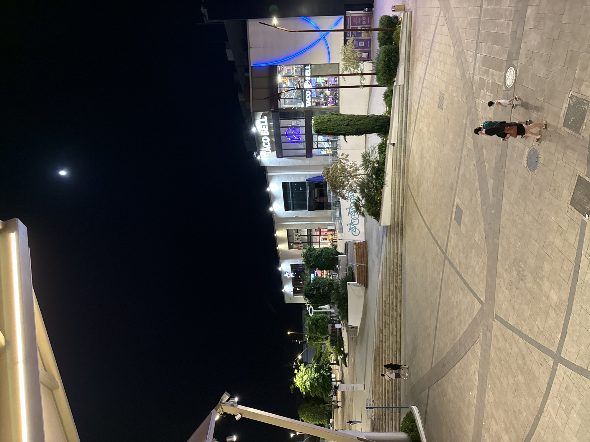 The media file depicts an outdoor scene at night, with a focus on a commercial building adorned with prominent "TACO BELL" signage, illuminated by bright blue neon lights forming a curved design. Adjacent to the Taco Bell is another establishment with a red star logo visible. The foreground features a paved walkway with geometric patterns, leading to a set of wide stairs. Several people are visible on the walkway, including a group of three people walking together, with one person carrying a bag, and another person walking ahead of them. There is also a lone figure on the stairs in the background. The sky above is dark, with the moon visible as a bright orb. The overall atmosphere suggests a lively urban environment at night, possibly a shopping or entertainment district in Alcorcón, Spain. The lighting from the businesses and streetlights creates a vibrant, yet slightly dramatic, ambiance.