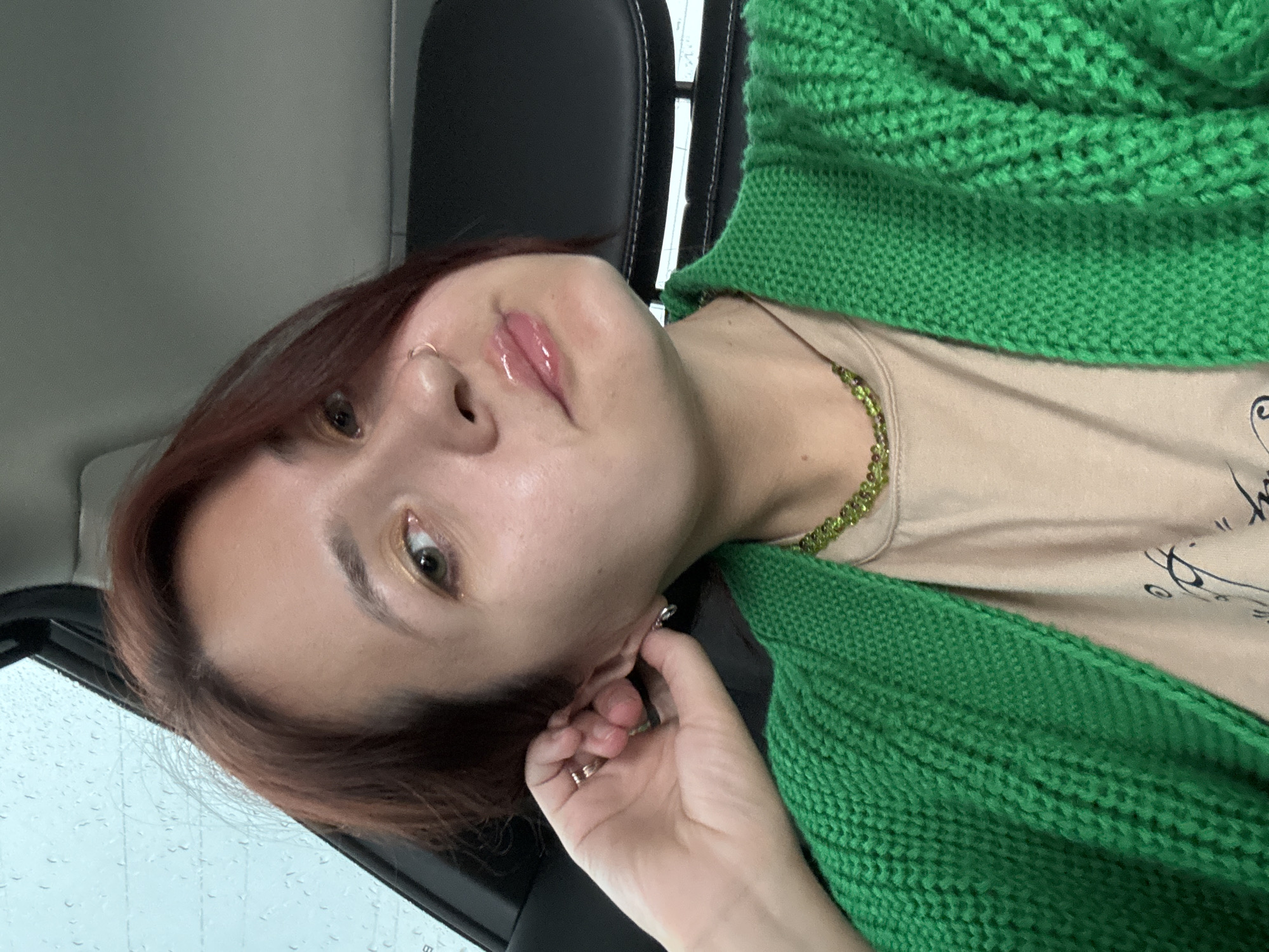 A woman with auburn hair and light skin is sitting in a car. She is wearing a bright green knitted cardigan over a beige t-shirt. A beaded green necklace is visible around her neck. Her right hand is raised to her ear, and she is wearing rings and an earring. She has a nose ring. Her lips are glossy. The background shows the interior of a car, with a black leather seat and a window with raindrops on it, suggesting it is raining outside. The location is the city of Naberezhnye Chelny, Russia.
