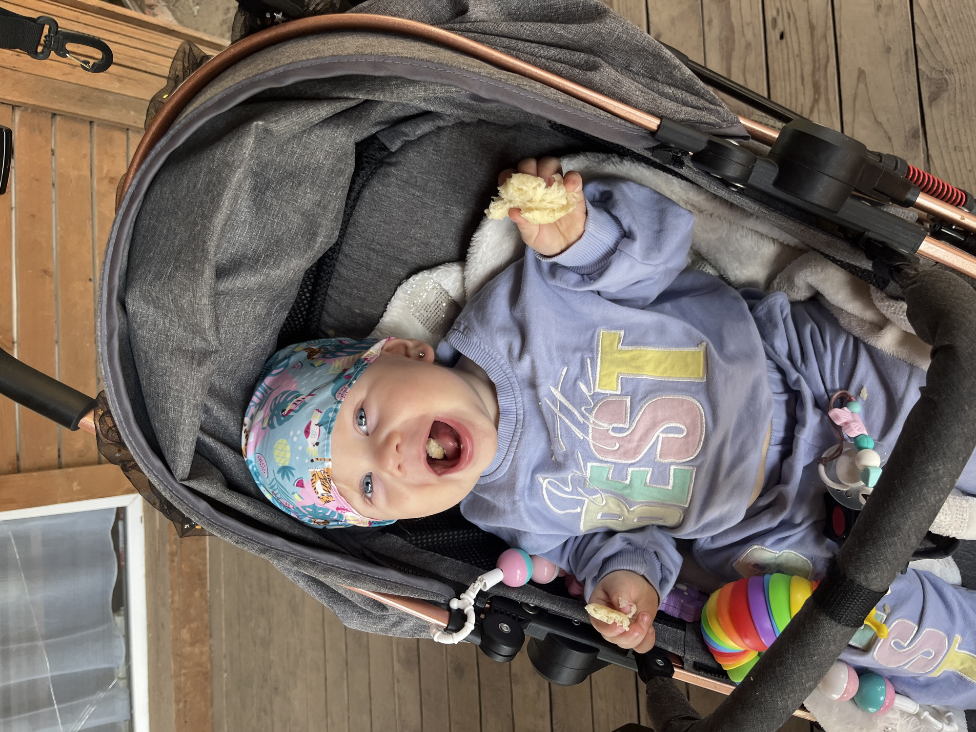 A baby girl sits in a stroller outdoors on a wooden deck. She is wearing a colorful headband and a light purple sweatshirt with the word "BEST" in pastel colors. Her mouth is open, and she is holding a piece of food in her right hand. The stroller is gray with rose gold accents. There are several toys attached to the stroller. The background shows a wooden structure with a window and curtains. The scene suggests a casual outdoor setting.
