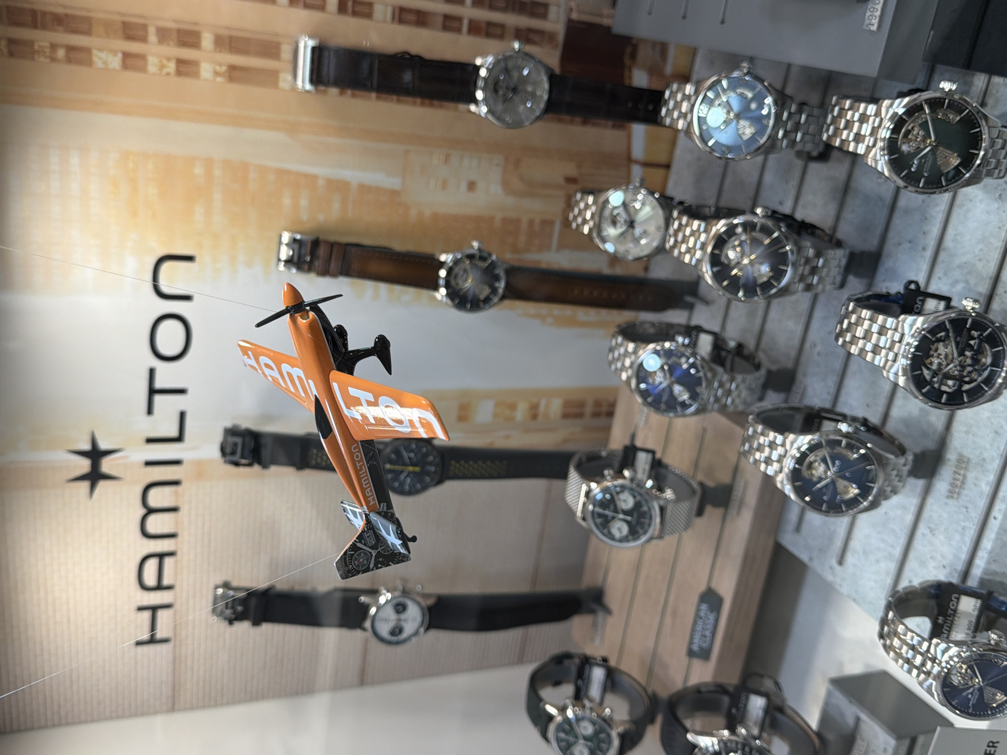 The image depicts a display case featuring a collection of Hamilton brand watches, alongside an orange toy airplane suspended by a thin wire. The backdrop behind the watches showcases the "HAMILTON" logo vertically. Various models of watches are arranged on wooden slats, showcasing different designs and materials, including leather and metal bands. The display is illuminated, and the overall setting suggests a retail environment. There is no explicit indication that the scene is located in Yerevan, Armenia.