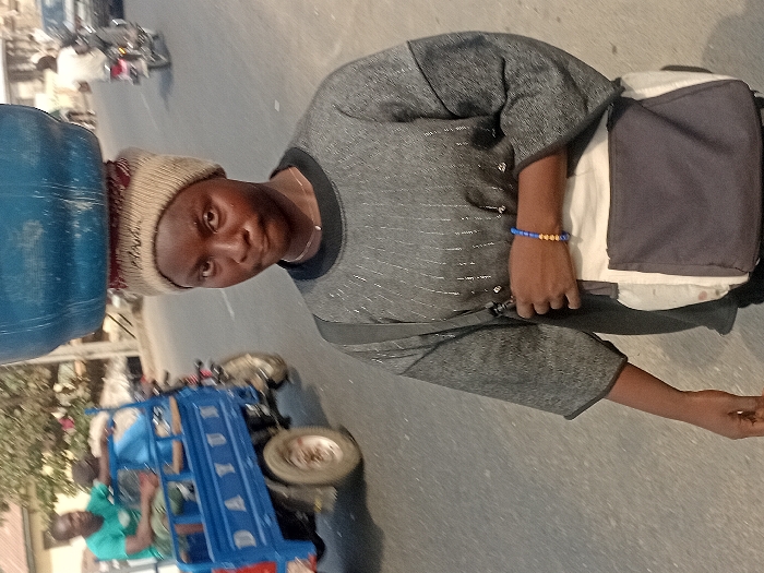 A person carries a large blue water jug on their head, wearing a beige knit hat and a gray, long-sleeved shirt. A messenger bag hangs from one shoulder. The person stands on what appears to be a road. In the background, a blue three-wheeled vehicle with two men in it is slightly blurred, along with other vehicles and indistinct buildings. The scene seems to be set in a bustling, possibly urban environment.
