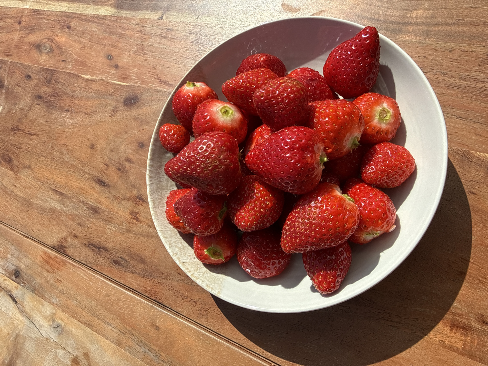 The image presents a close-up, high-angle view of a white, shallow bowl overflowing with fresh, ripe strawberries. The bowl is positioned in the lower right section of the frame, resting on a rustic wooden table.
The strawberries are the primary subjects, appearing in various sizes but uniformly vibrant red, with some showcasing lighter red or pinkish areas, and distinct yellow seeds (achenes) dotting their surfaces. Several strawberries still have small remnants of their green calyx or the pale green core where the stem was attached. The fruit appears plump and slightly glossy, suggesting freshness.
The bowl itself is simple and round, made of a light grey or off-white material with a subtle, textured finish. It casts a prominent, well-defined shadow across the wooden surface, indicating bright, direct sunlight.
The wooden table, serving as the setting, is constructed from wide planks. It features a warm, reddish-brown hue with visible wood grain, knots, and natural imperfections, adding to the rustic aesthetic. The strong, crisp shadows cast by the bowl and individual strawberries, along with bright highlights on the fruit, suggest the photo was taken in clear, sunny weather, likely during the late morning or early afternoon. This outdoor-like illumination, possibly on a balcony or patio in Sofia, Bulgaria, highlights the natural vibrancy of the scene. No people or visible text are present. The overall impression is one of natural abundance, freshness, and the simple beauty of ripe fruit.
