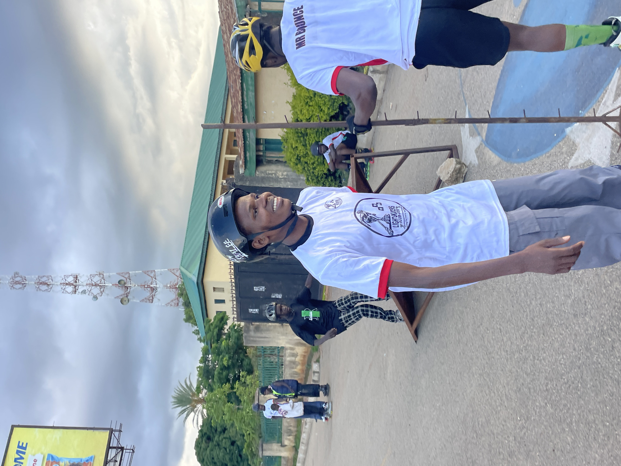 In Jos, Nigeria, under a cloudy sky, a group of young people engage in rollerblading. The foreground features two individuals wearing helmets and rollerblading gear. One, wearing a white t-shirt with a red trim and grey shorts, smiles at the camera. The other, in a white t-shirt with "MR BOUNCE" printed on the back and black shorts, faces away. In the mid-ground, a person in a black t-shirt and checkered pants practices on a ramp. Further back, two individuals in casual wear are visible, one in a white t-shirt and the other in a blue vest. The environment includes a large communication tower, a billboard with the text "ME," a building with a green roof, and residential fencing, indicating an urban setting. The ground is paved, with a blue painted area visible to the right.