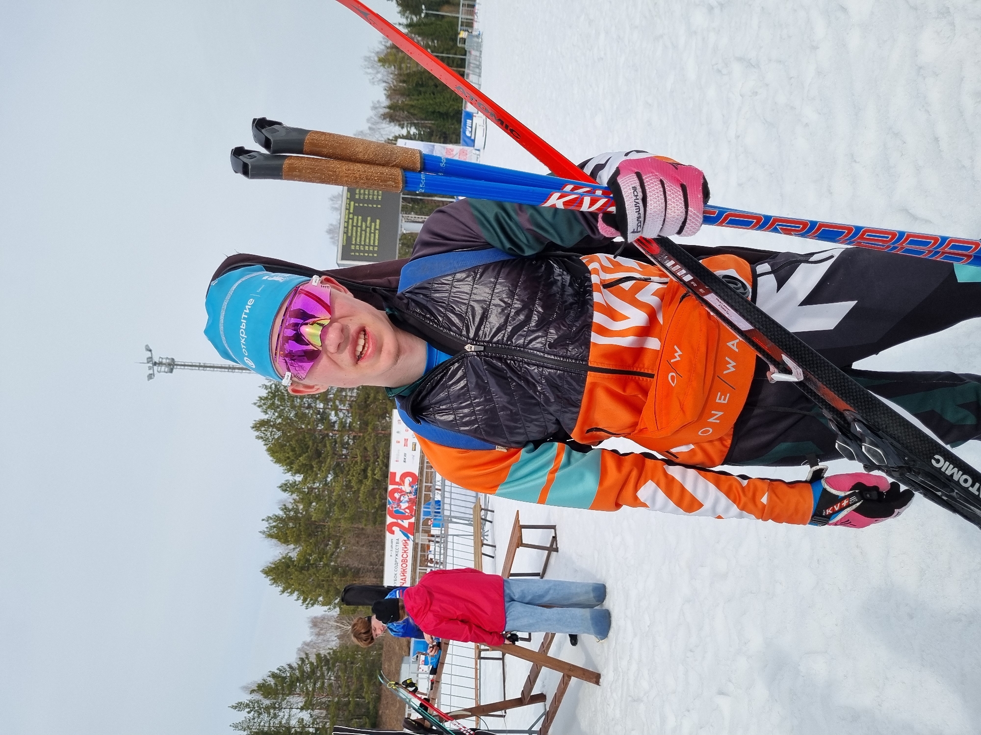 The image displays an outdoor winter sports environment within the Прикамский region of Russia, specifically a ski track, referencing Chaykovsky on a background banner.
In the foreground, a young male stands facing the camera. He wears a blue beanie with an "открытие" logo, pink reflective ski goggles, and a clear nose guard. His attire includes a black quilted vest over an orange, teal, and black long-sleeved top, black and white ski pants, pink and black gloves (one bearing "БОЛЬШУНОВ"), and an orange fanny pack with "ONE WAY" lettering. He holds a pair of red and black "ATOMIC" skis with bindings in his left arm/hand and two blue and red ski poles with cork grips in his right hand. His mouth is slightly ajar.
In the midground, two other individuals are present on the snow-covered ground. One person, wearing a red hooded jacket and light blue pants, stands with their back to the camera near a wooden bench. Another individual in a blue top is partially obscured behind a wooden ski rack holding several pairs of skis.
The background features a line of coniferous trees beneath an overcast sky. A tall metal light tower is visible on the right. A large white banner with red text, reading "КУБОК СОДРУЖЕСТВА ЧАЙКОВСКИЙ 2023" and "2023," is displayed behind metal barriers. To the right of the banner, a digital scoreboard shows numerical data and the "открытие" logo. The ground surface throughout the scene is covered in snow.