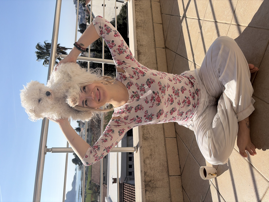 A female subject is seated cross-legged on a light-tan tiled outdoor balcony or terrace in Budva, Montenegro. She wears a white long-sleeved shirt with a pink floral pattern and light-colored striped pants. A small, white, fluffy dog is positioned on top of her head, held by her raised hands. The dog faces forward with its eyes partially closed. The woman exhibits a slight smile and looks towards the left. Her left wrist displays a dark beaded bracelet, and a silver-colored necklace with a circular pendant is visible. A white ceramic mug with a gold handle, containing a dark liquid, rests on the tiled floor to her left. The background features a silver-colored metal railing, behind which lies a panoramic view of Budva, including residential buildings with red-tiled roofs, green vegetation, a palm tree on the right, and the Adriatic Sea with a distant landmass on the left. The sky is clear blue, and bright sunlight casts distinct shadows on the subjects and the tiled surface.