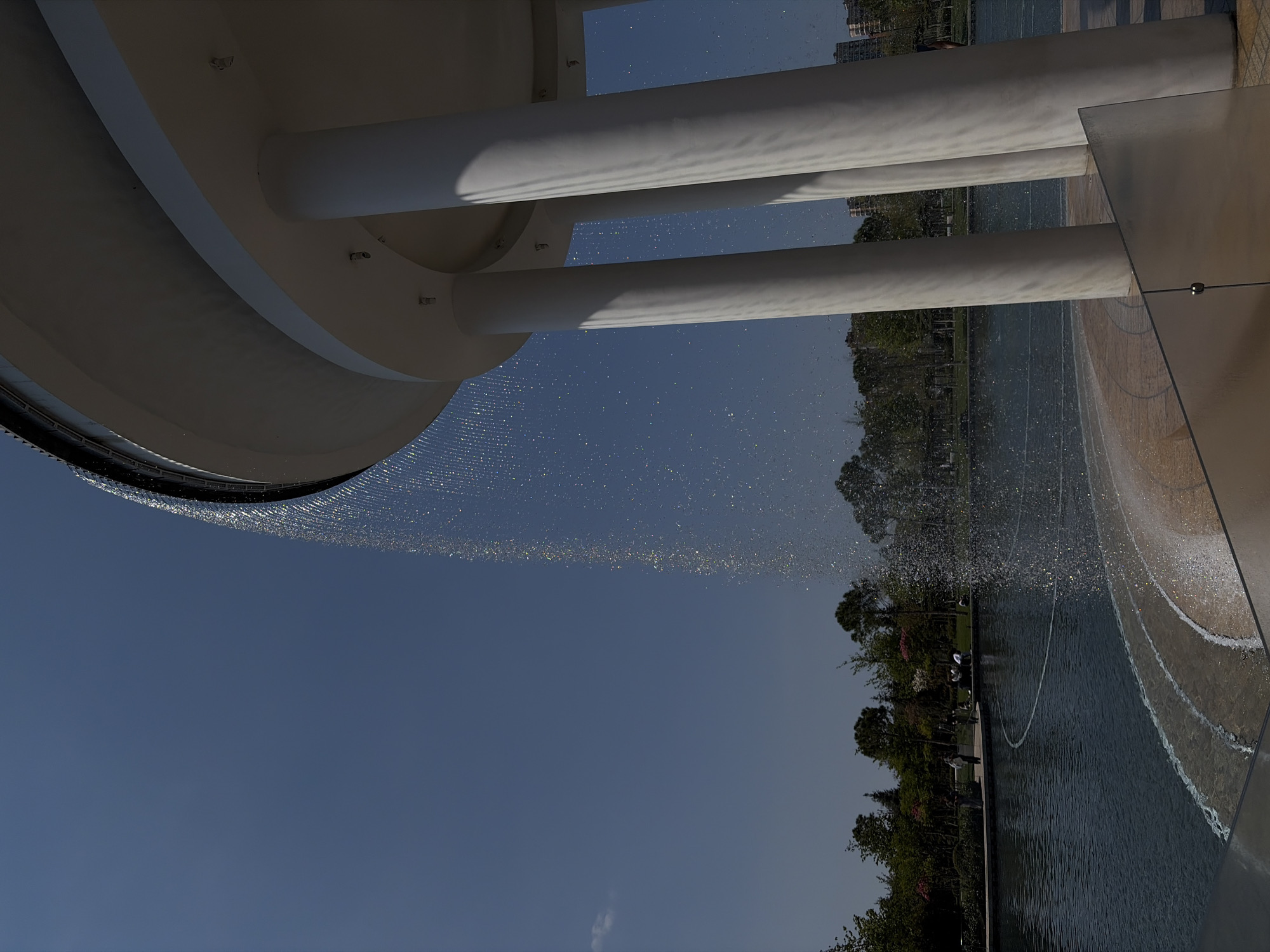 A low-angle view captures a modern water fountain installation in Krasnodar, Russia. The structure consists of a broad, white circular canopy supported by several white cylindrical pillars. Water cascades from the canopy's edge, creating a shimmering curtain effect. The water collects in a dark pool below, where concentric circles are visible on the surface.
In the background, trees and manicured parkland extend towards the horizon, suggesting an urban park setting. A pathway runs alongside the pool, with a few people visible along its edge. The sky above is a clear, pale blue. A geometric, reflective structure is located on the right.