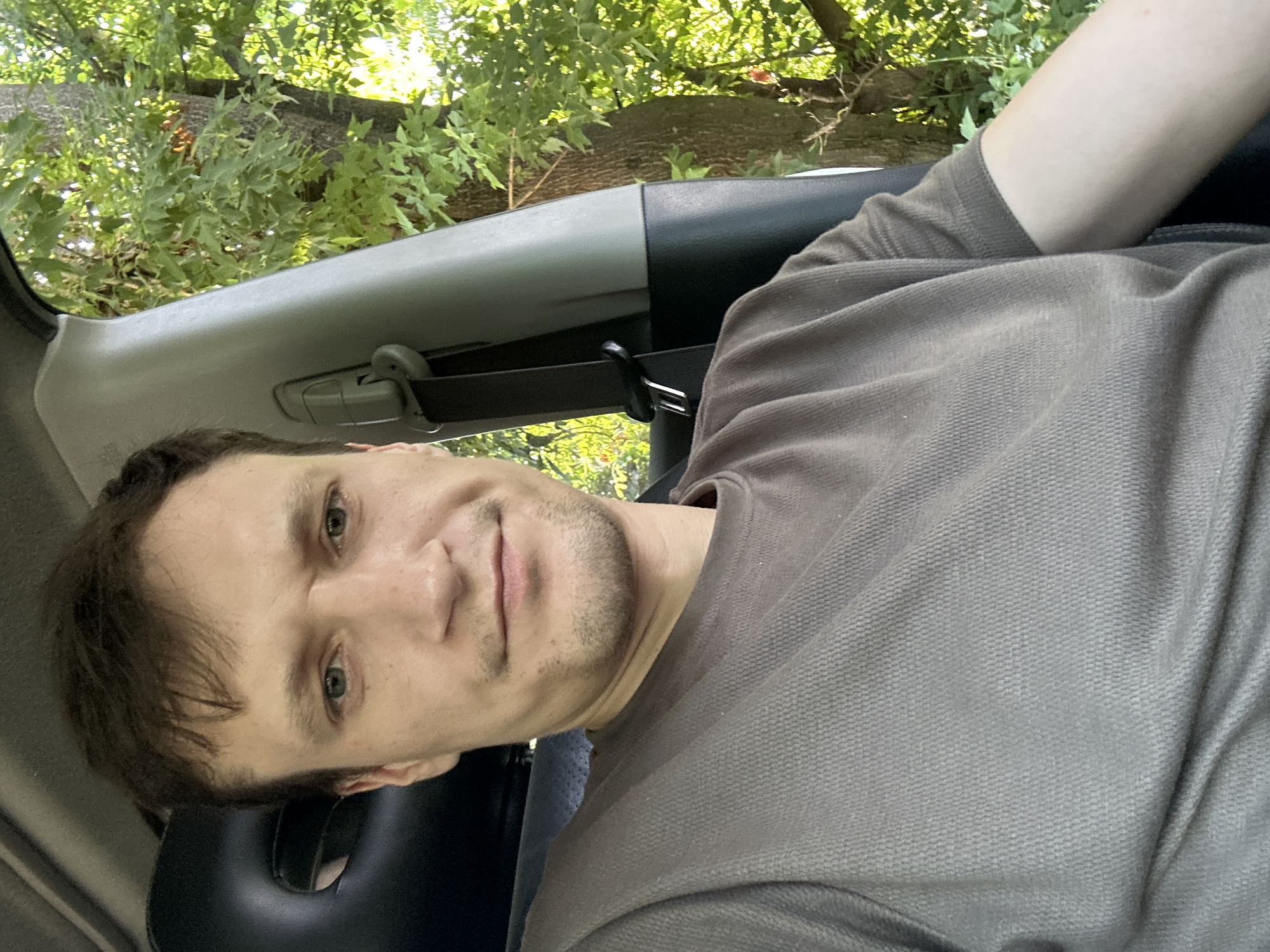 A selfie captured from the perspective of someone sitting in the driver's seat of a vehicle. The individual, a light-skinned male with short brown hair and light green eyes, is wearing a dark grey, short-sleeved t-shirt. He has a slight stubble on his chin and upper lip. The interior of the car is visible, including a black seat with a headrest and a grey pillar with a seatbelt buckle. Through the car window, a lush green environment with trees and foliage is visible, bathed in sunlight, suggesting an outdoor setting, possibly on the outskirts of Makiv, Ukraine.