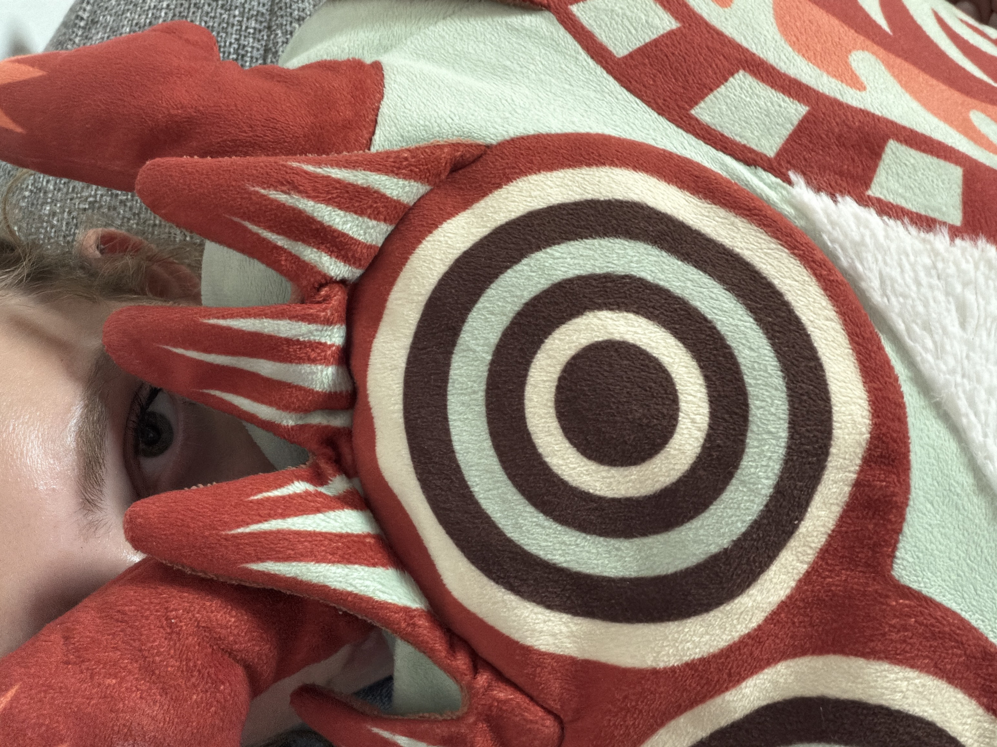 A close-up shot captures a person's eye peeking through a red, plush toy that resembles a dragon. The toy has a circular pattern with concentric rings of red, cream, and brown, as well as sharp, red and white striped fins. The person's eye is dark brown with a hint of green and framed by an eyebrow. The background consists of a grey, textured surface, possibly a blanket or couch. The image does not provide any specific details about the city of Ashkelon, Israel, or any identifiable landmarks within the city.