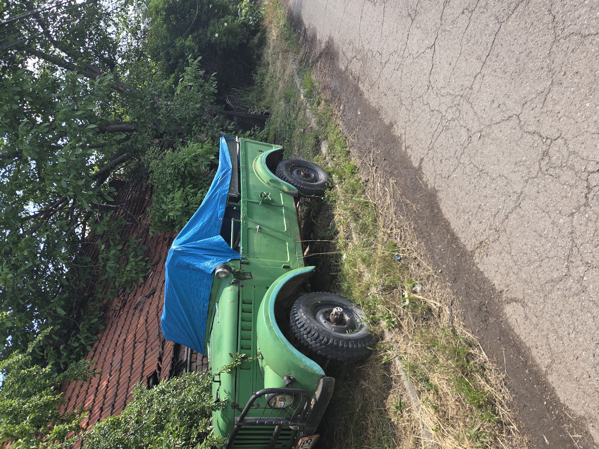 The image displays an outdoor scene in Elin Pelin, Bulgaria, on a clear or partly cloudy day. The primary subject is a faded green, possibly military-style, utility vehicle parked on an overgrown grass and dirt verge beside a cracked asphalt road. No individuals are visible.
The vehicle, appearing to be an older model 4x4, features a robust design. Its front left section is prominently visible, showing a round headlight, a black tubular grille guard, and a fragment of a white license plate with black characters, "OCB" being legible. The vehicle's paint is a worn green, with hints of a lighter blue paint present on the front left fender above the wheel. The front left wheel is equipped with a chunky, off-road tire. The open-top rear section and parts of the cabin are covered by a wrinkled, bright blue tarpaulin. Green plant tendrils are seen growing over the vehicle's front fender.
Behind the vehicle, an old building with a reddish-brown tiled roof is partially obscured by dense green foliage. The terracotta roof tiles exhibit wear and damage, with some appearing broken or missing. Large, mature deciduous trees with lush green leaves dominate the background and upper sections of the image, contributing to the overgrown appearance of the area surrounding the building.
The ground where the vehicle is parked consists of dry, yellowish grass mixed with green weeds and exposed soil. A distinct, uneven edge separates this unkempt verge from the adjacent road. The road surface is asphalt, severely weathered with a network of cracks and fissures across its visible expanse. The lighting suggests direct sunlight, creating subtle shadows.