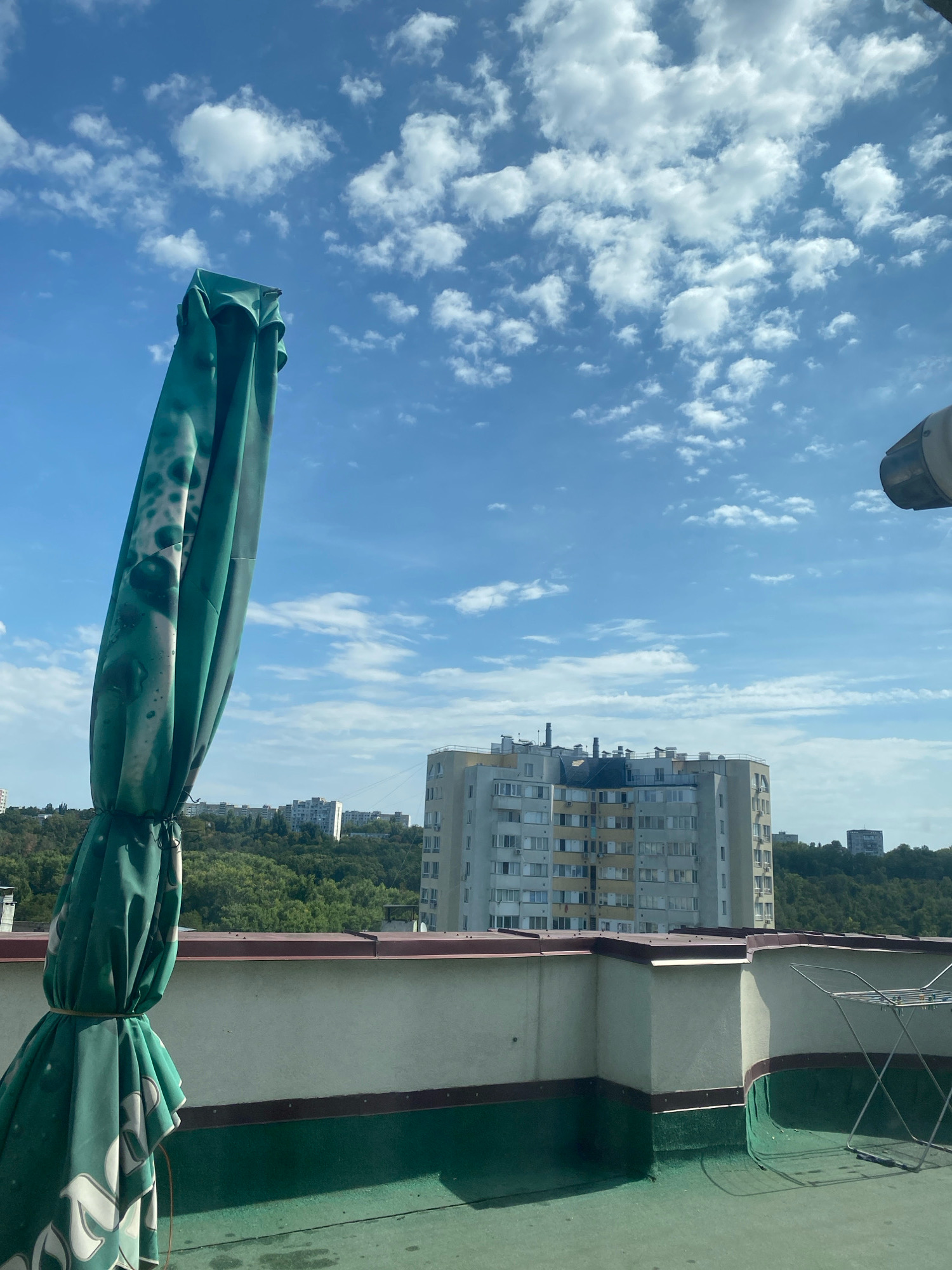 An outdoor rooftop scene in Chișinău, Moldova, features a folded green umbrella in the foreground, tied with string. A light green rooftop surface extends into the mid-ground, bordered by a white wall with a dark red trim. In the background, a densely wooded hill slopes upwards, with several multi-story apartment buildings visible amongst the trees and on the horizon. The sky is a clear blue with scattered white clouds. A drying rack is present on the rooftop to the right.