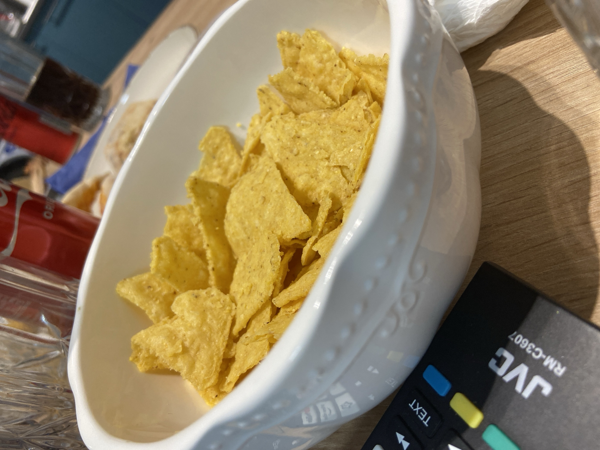 A close-up, slightly overhead photograph captures a casual snack arrangement set on a light wooden table, likely indoors in Sofia, Bulgaria. The primary subject is a decorative white serving dish, generously filled with golden-yellow tortilla chips, commonly known as nachos. The chips are lightly seasoned, with small dark flecks visible on their surface.
To the left of the serving dish, a red Coca-Cola can is partially visible, clearly displaying the brand name "Coca-Cola" and the text "ORIG" (likely part of "ORIGINAL TASTE"). Adjacent to the can is a clear glass holding a dark beverage, presumably cola, with ice.
On the right side of the frame, a black JVC brand remote control is positioned on the table. The brand "JVC" and model number "RM-C3607" are prominently visible, along with the word "TEXT" on one of its functional buttons. Other buttons show blue, yellow, and turquoise colored accents. Behind the main dish, a crumpled white napkin or paper towel is also present on the wooden surface.
Bright natural light illuminates the scene from the left, creating distinct shadows from the serving dish and the remote control on the table, indicating it is daytime. The overall ambiance suggests a relaxed activity, such as enjoying snacks and a drink, possibly while watching television. No people are visible in the image.