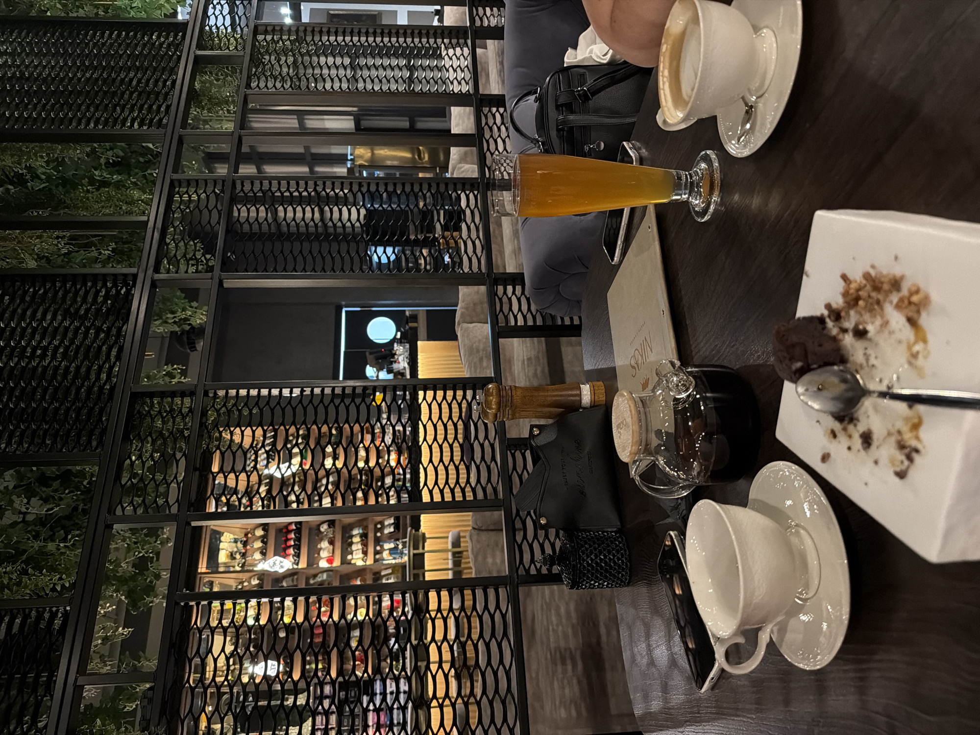 The image captures an indoor scene in Krasnoyarsk, Russia, possibly a restaurant or café. The setting includes a table with several items: two teacups, one containing a beverage with a creamy layer, a glass of amber-colored liquid (possibly beer), a white square plate with remains of a dessert (brownie and toppings), a spoon, a menu, a small teapot, and a black purse. A person is partially visible, seated at the table. The background features a decorative metal grid structure with plants, partially obscuring shelves stocked with bottles, presumably wine. The room is illuminated, creating a warm, inviting atmosphere.