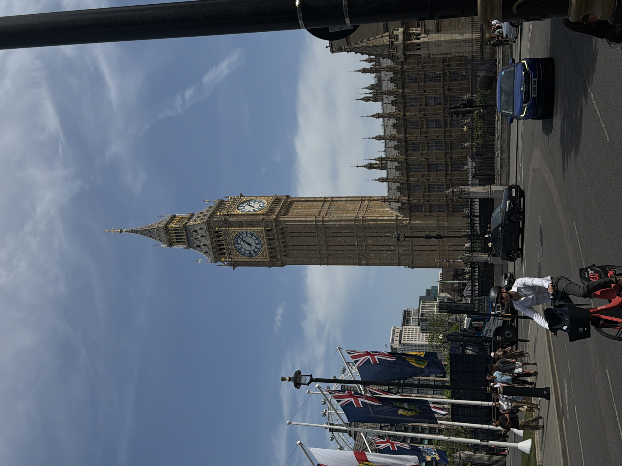 The image captures a scene in London, United Kingdom, featuring the iconic Big Ben and the Houses of Parliament. The sky is blue with sparse clouds. British flags are displayed prominently in the foreground. A group of people is visible on the sidewalk, and a man is riding a red bicycle in the street. Cars are driving on the road alongside pedestrians.